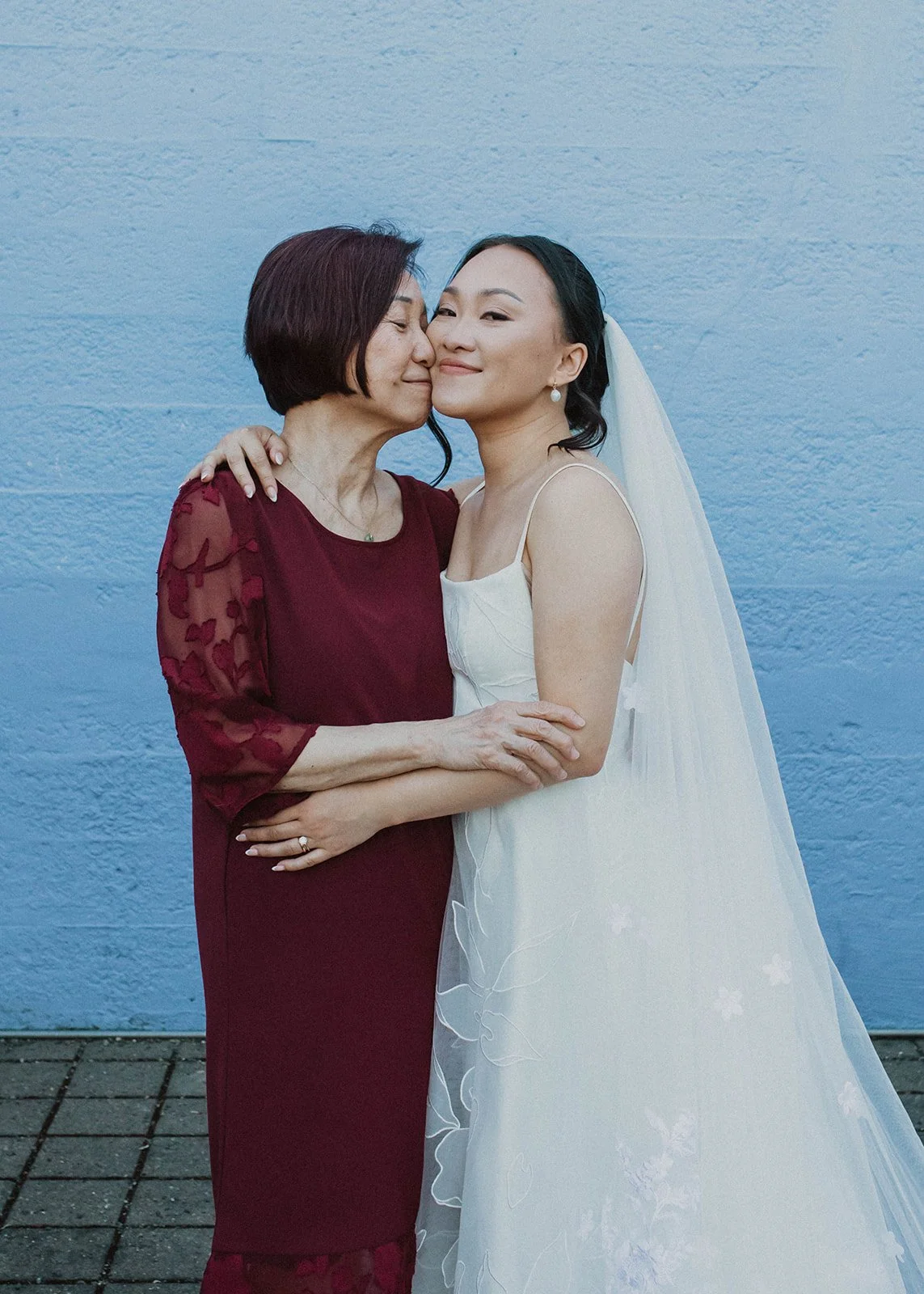 Bride shares a close portrait with her mother following a Portland wedding ceremony, standing against a blue outdoor backdrop.