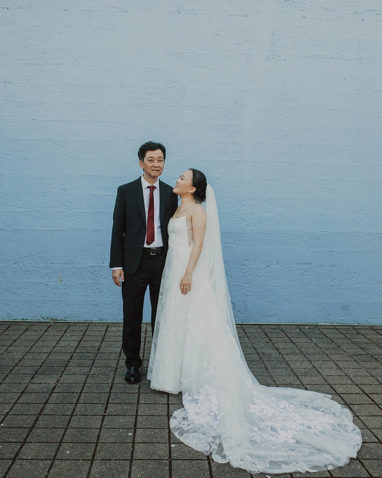 Bride stands beside her father for a wedding portrait after the ceremony, photographed against a blue outdoor backdrop in Portland.