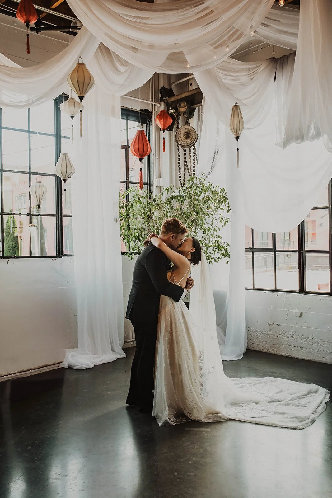 Newly married couple shares their first kiss beneath suspended drapery and lanterns during a modern Portland wedding ceremony.