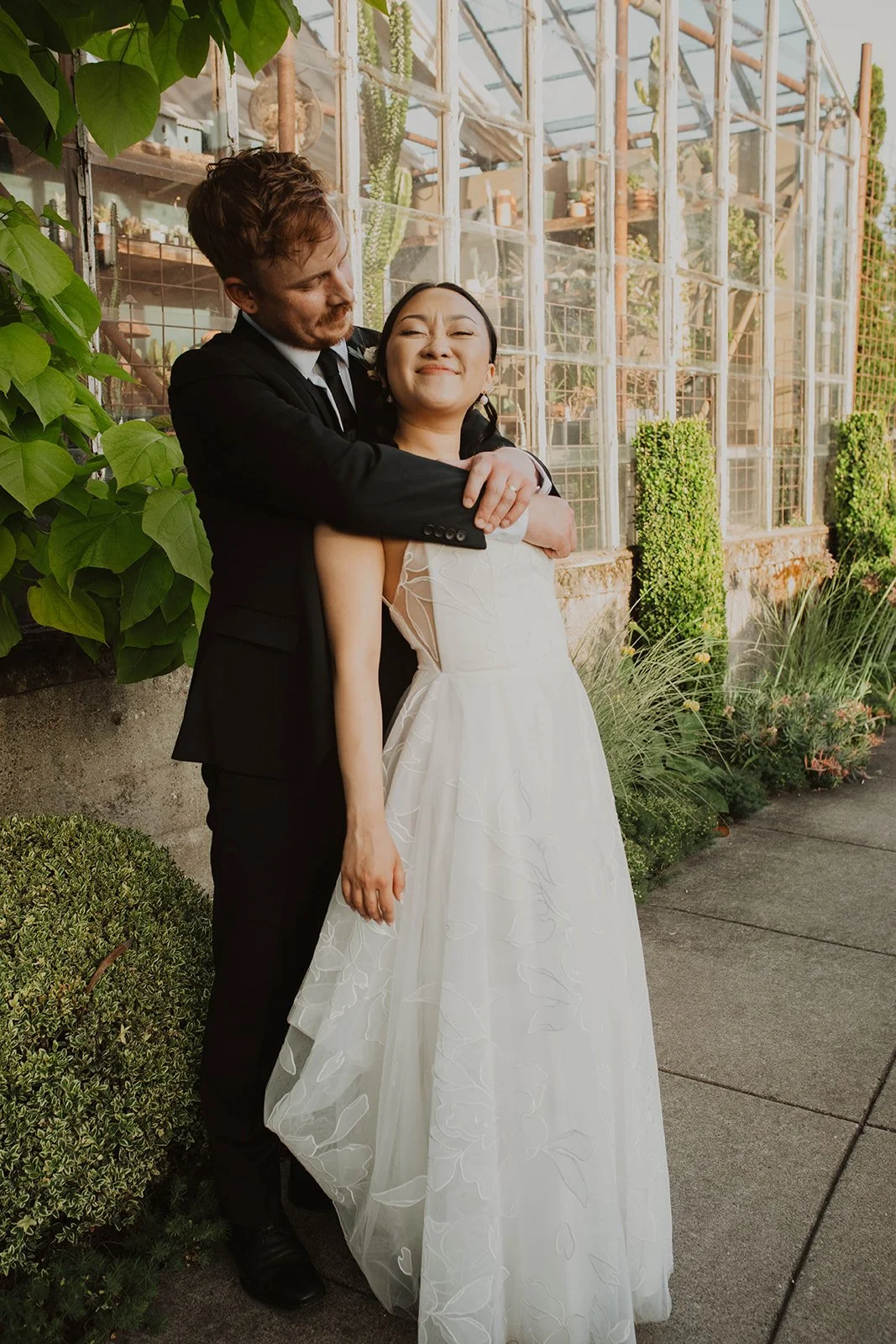 Groom wraps his arms around his bride as they smile together during intimate wedding portraits beside a greenhouse and garden pathway.