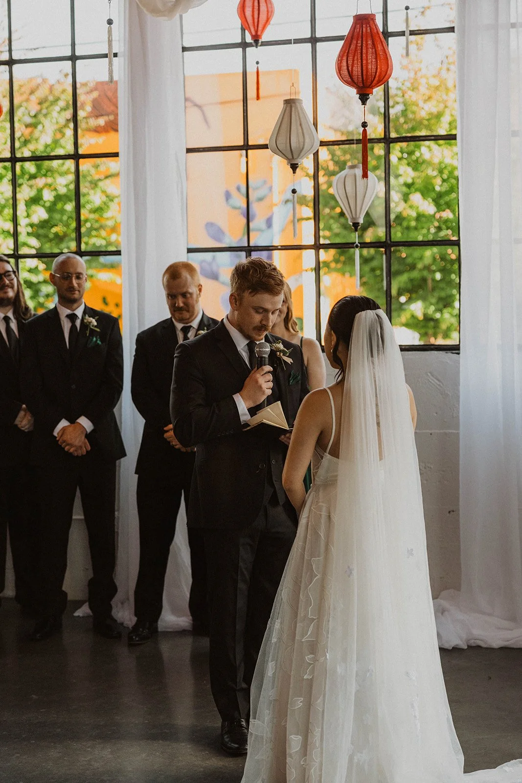 Groom reads handwritten wedding vows during an emotional ceremony at a modern Portland wedding venue with lantern and drapery decor.