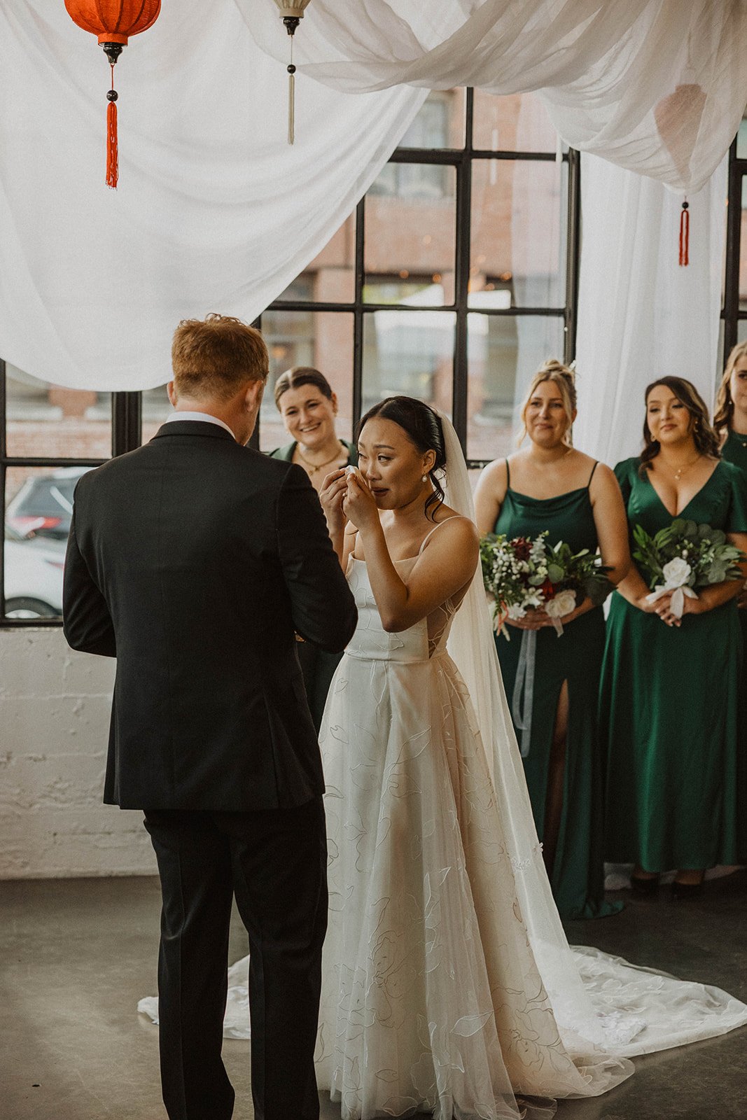 Bride wipes away tears while listening to wedding vows during an emotional ceremony at a Portland wedding venue with industrial windows and draped fabric decor.
