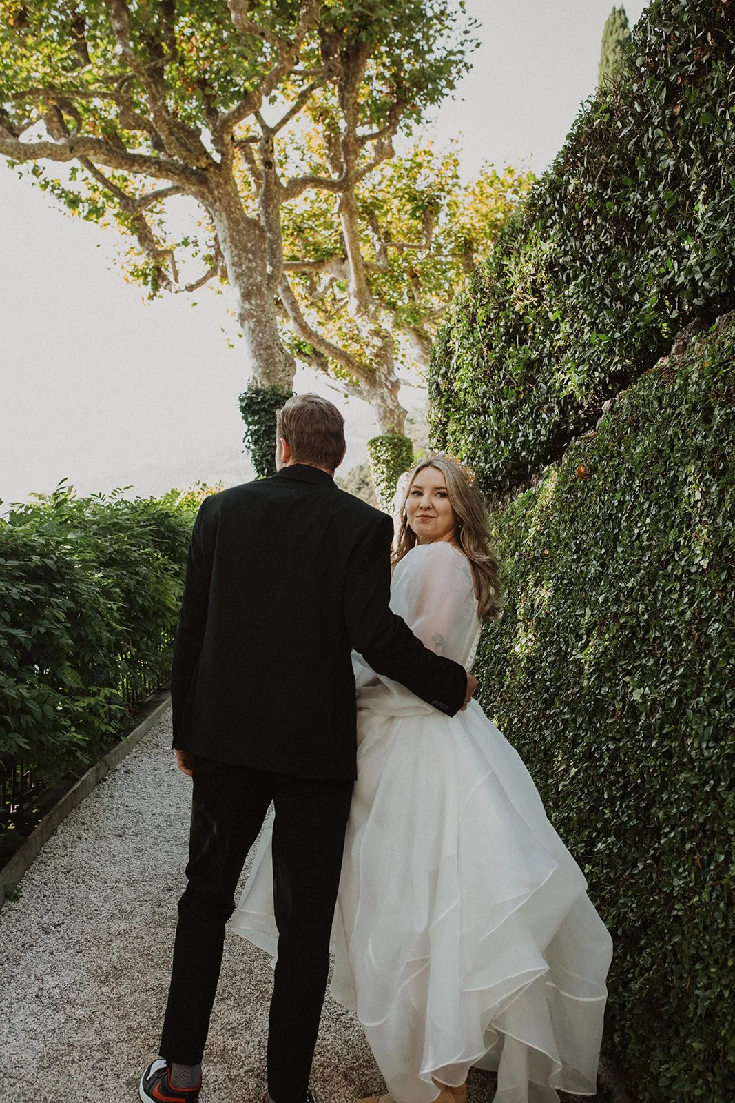 Bride looks back toward the camera while walking with her groom through the garden paths at Villa del Balbianello on Lake Como.
