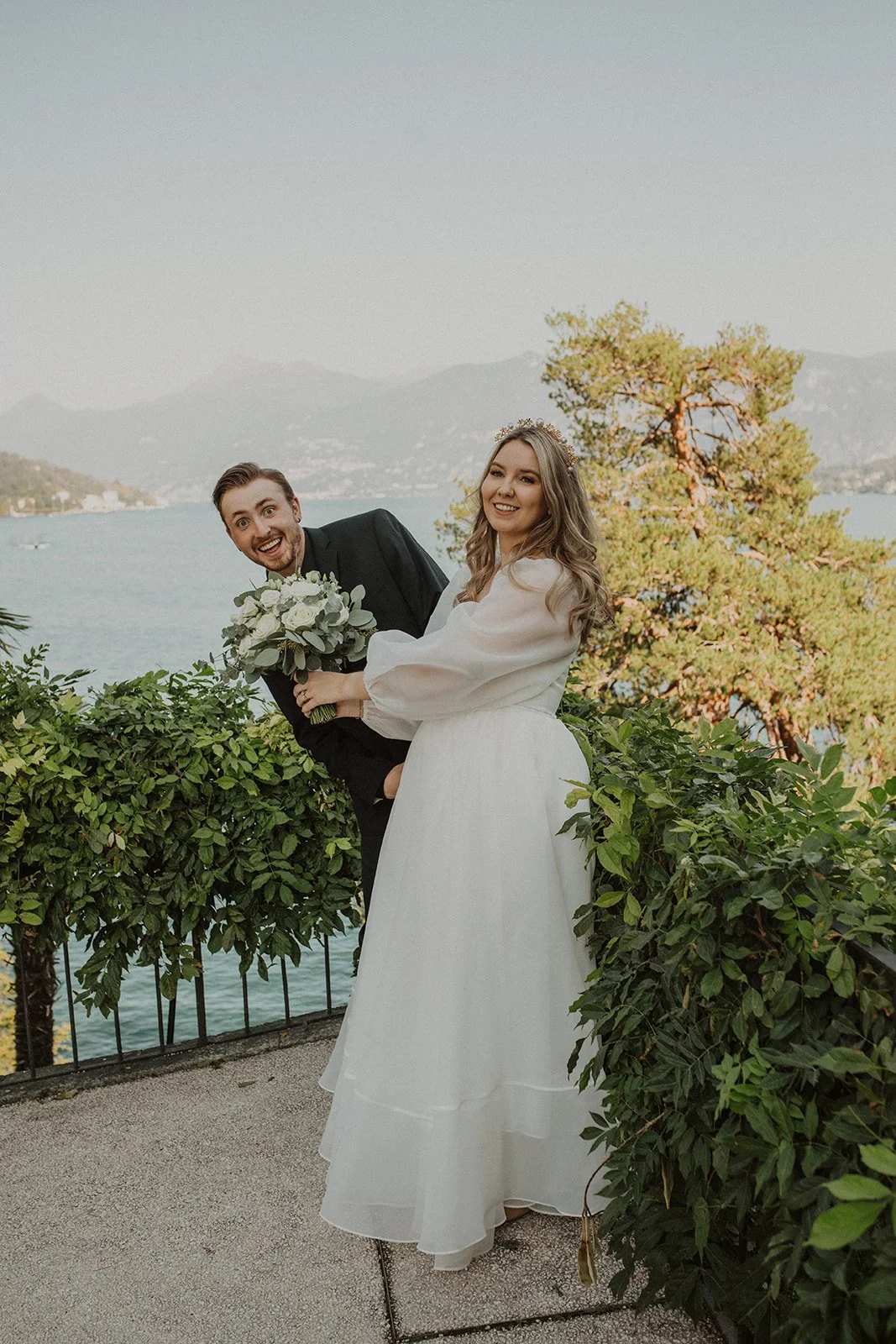 Bride smiling while the groom playfully peeks around her during a fun portrait overlooking Lake Como.
