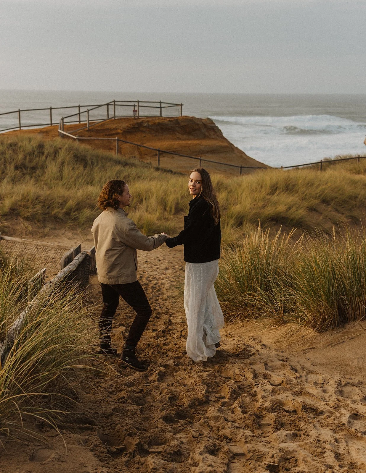 Woman looks back at fiance while walking through the coastal dunes at Cape Kiwanda during their engagement session.