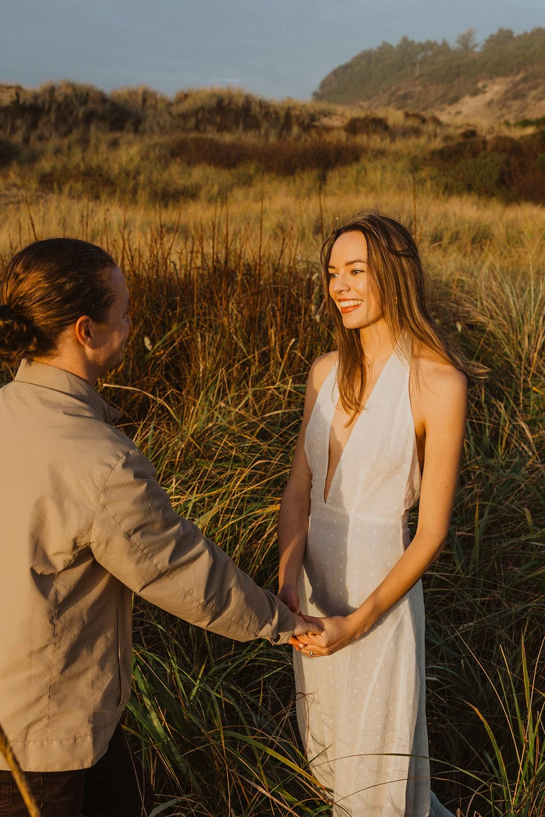 Couple hold hands in the coastal grasses at Cape Kiwanda as golden evening light fills their Oregon Coast engagement session