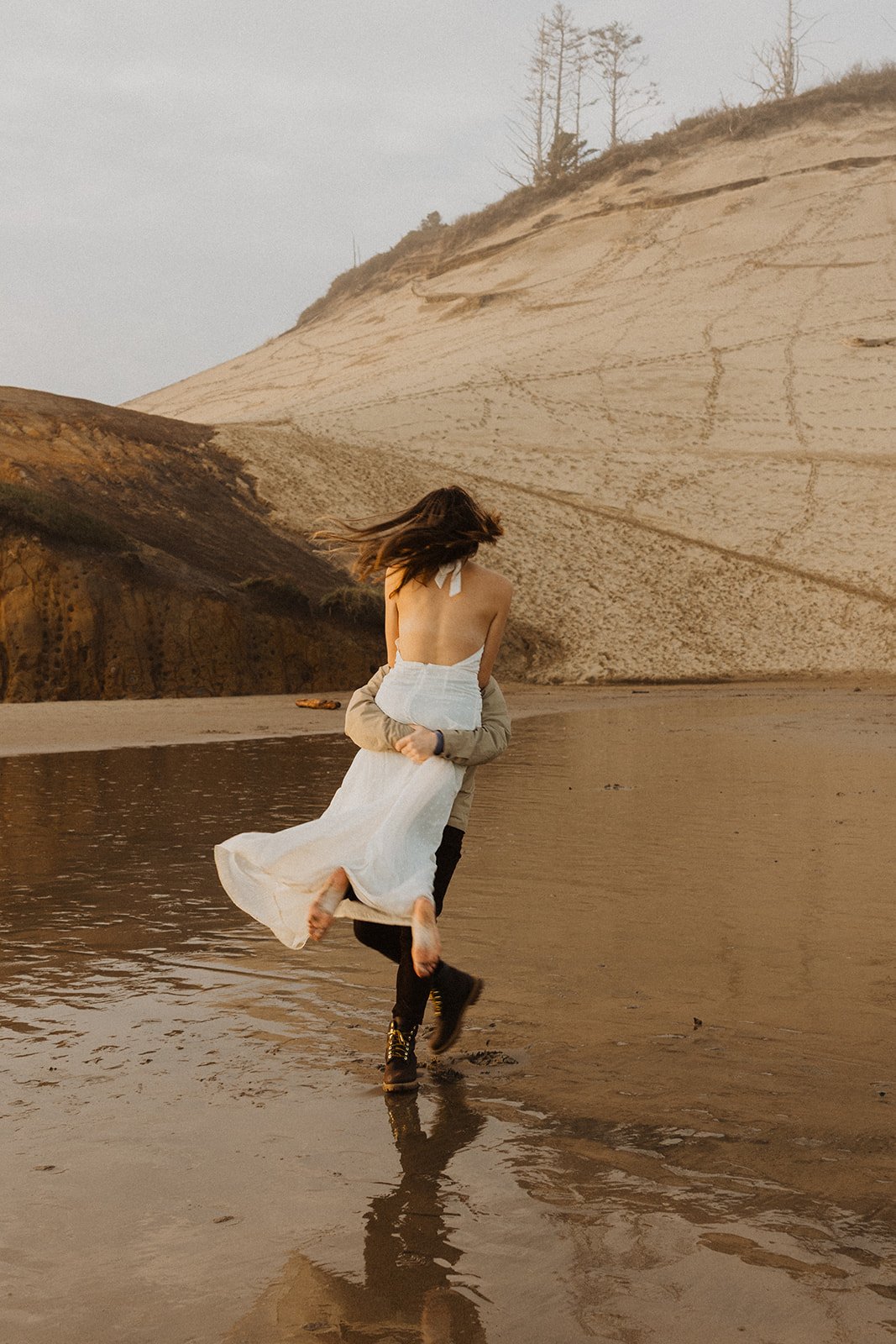 Lucas carries Miranda along the wet sand beneath the cliffs during their Cape Kiwanda engagement session