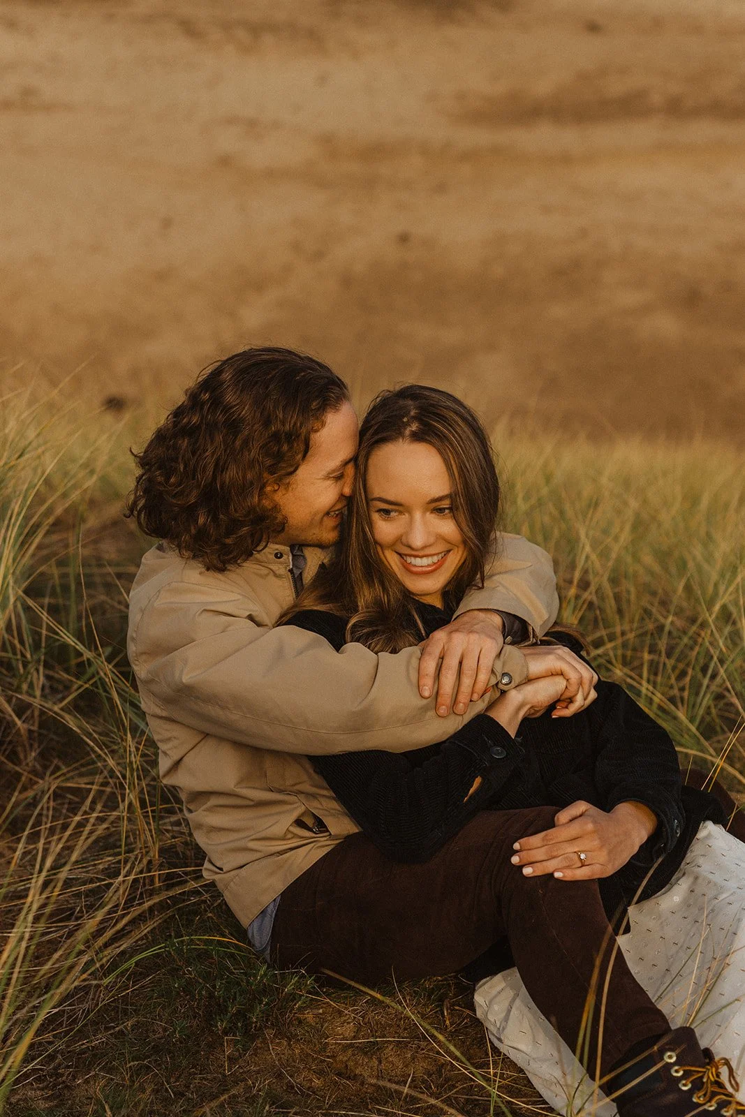Couple snuggle in the coastal grasses at Cape Kiwanda as golden evening light fills their Oregon Coast engagement session