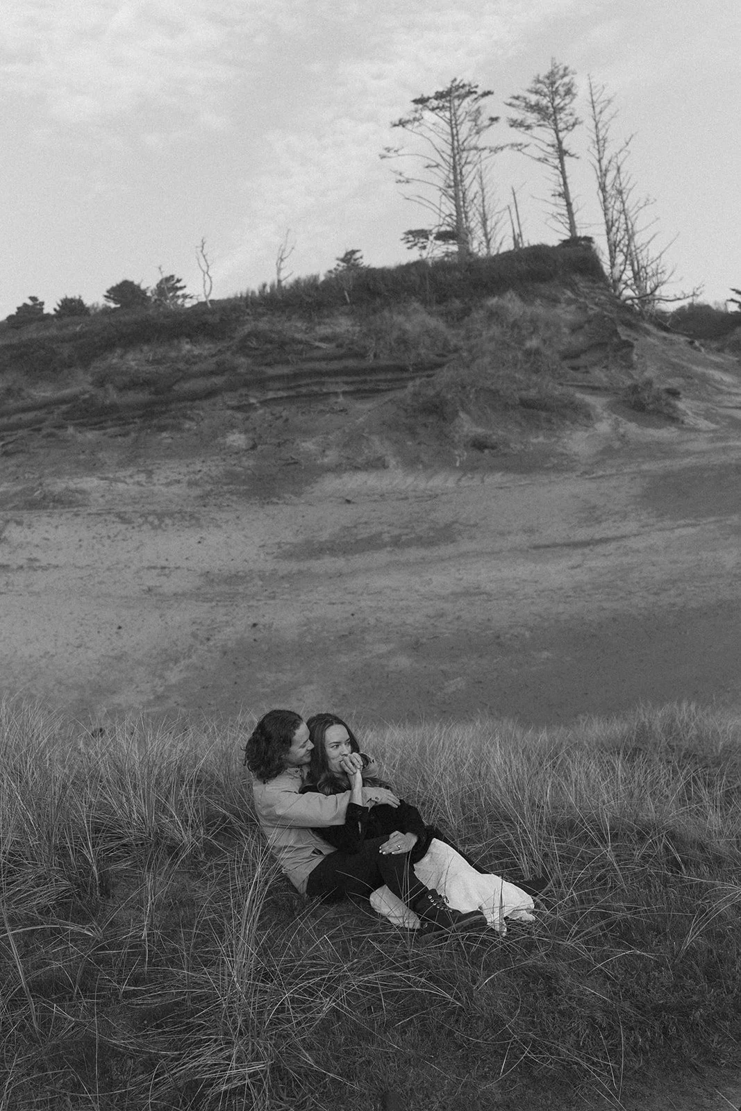 Couple sit in the tall grass with dunes in the distance during their Pacific City engagement session