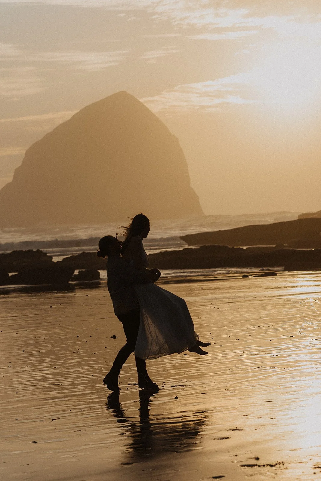 Lucas spins Miranda while they laugh together in the surf during their playful Cape Kiwanda engagement session on the Oregon Coast
