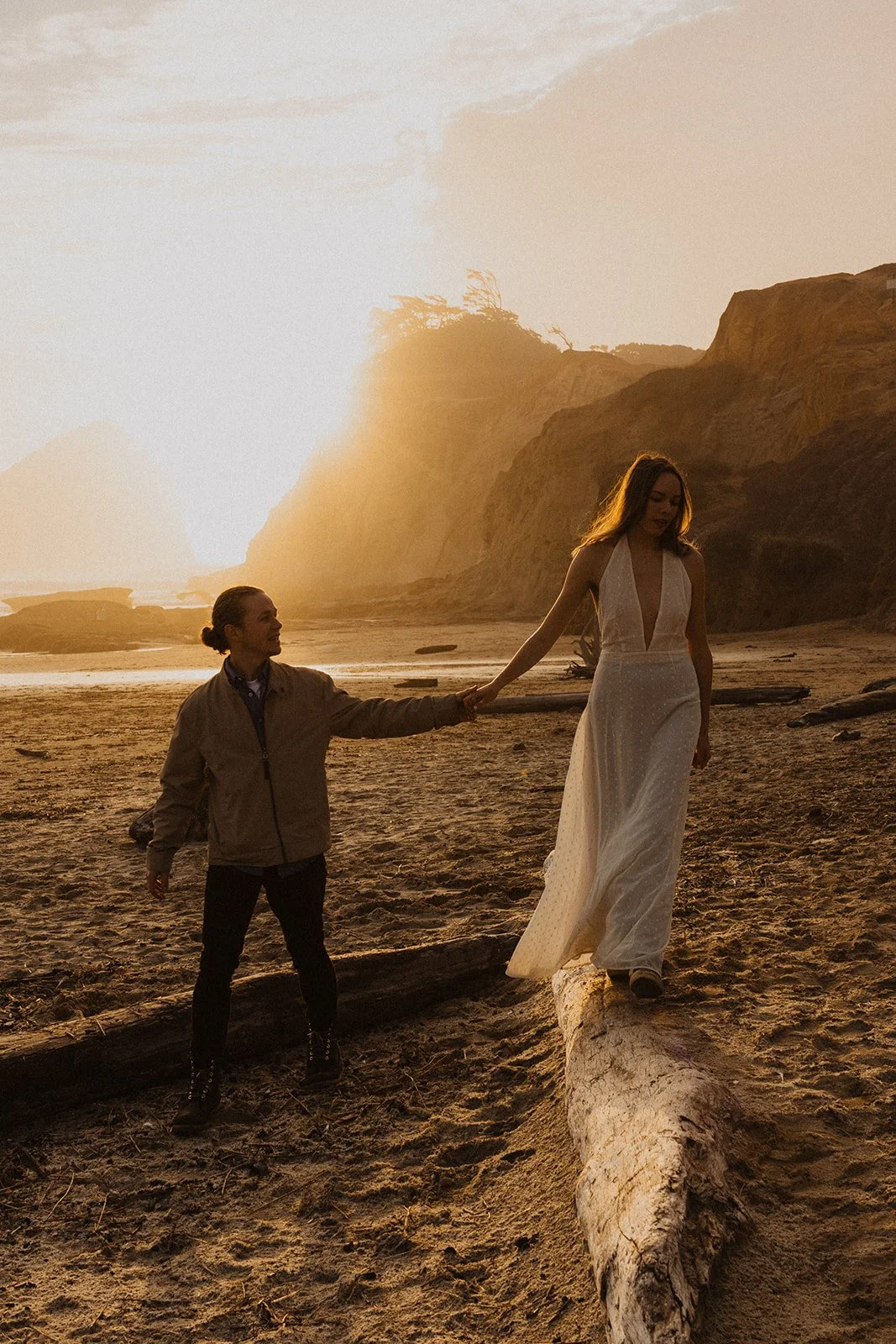 Miranda carefully walks across a driftwood log while holding Lucas's hand during their Cape Kiwanda engagement session on the Oregon Coast