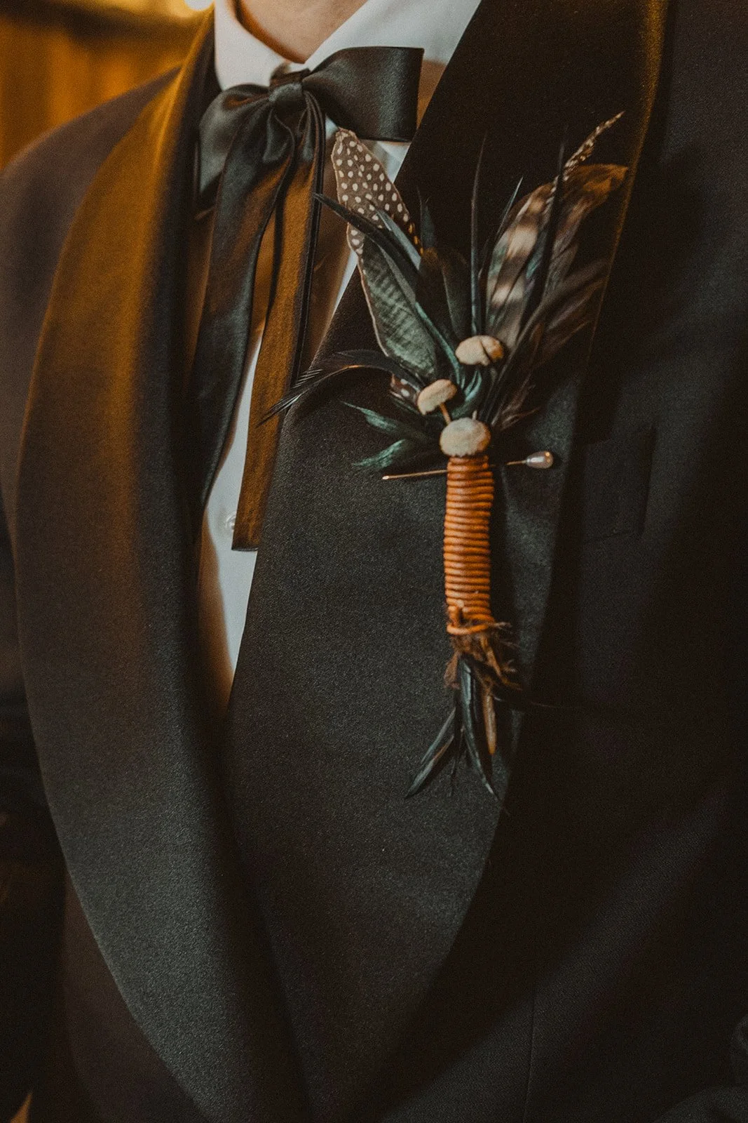 Close-up of the groom's black tuxedo with a ribbon bow tie and feather boutonniere at Bright Morning Farm.