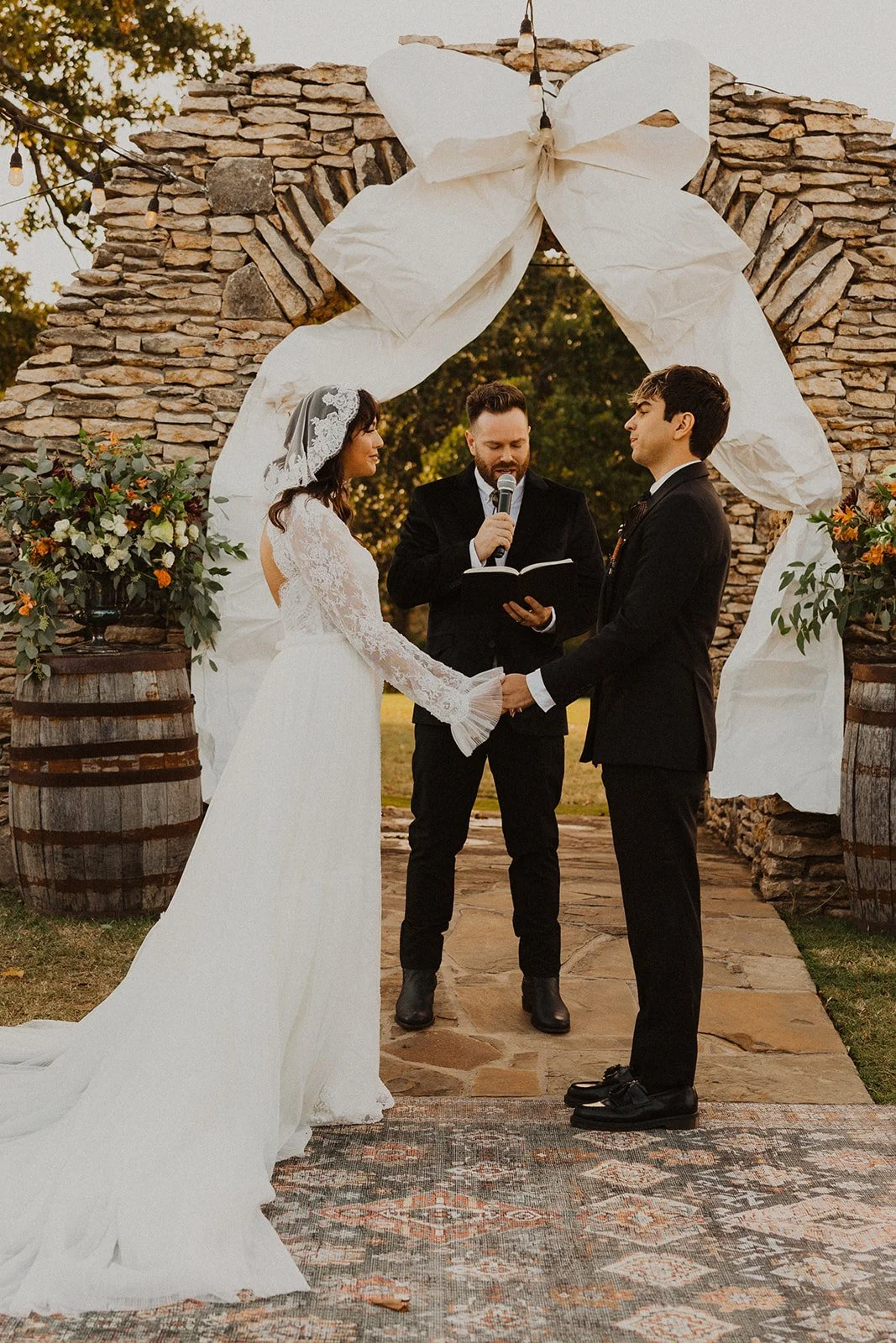 Bride and groom holding hands during their outdoor wedding ceremony beneath the stone arch at Bright Morning Farm as the officiant speaks.