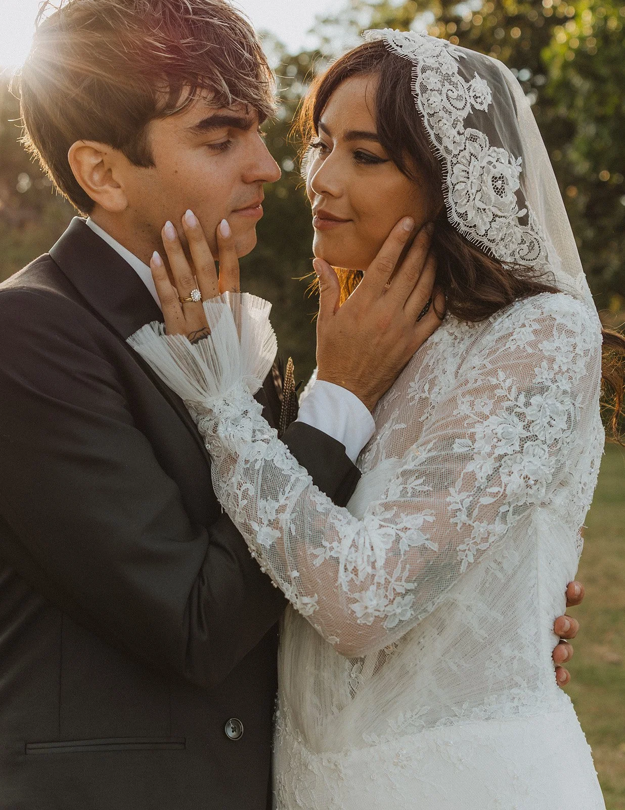 Golden hour wedding portrait of the bride and groom embracing, highlighting the bride's lace veil and engagement ring.