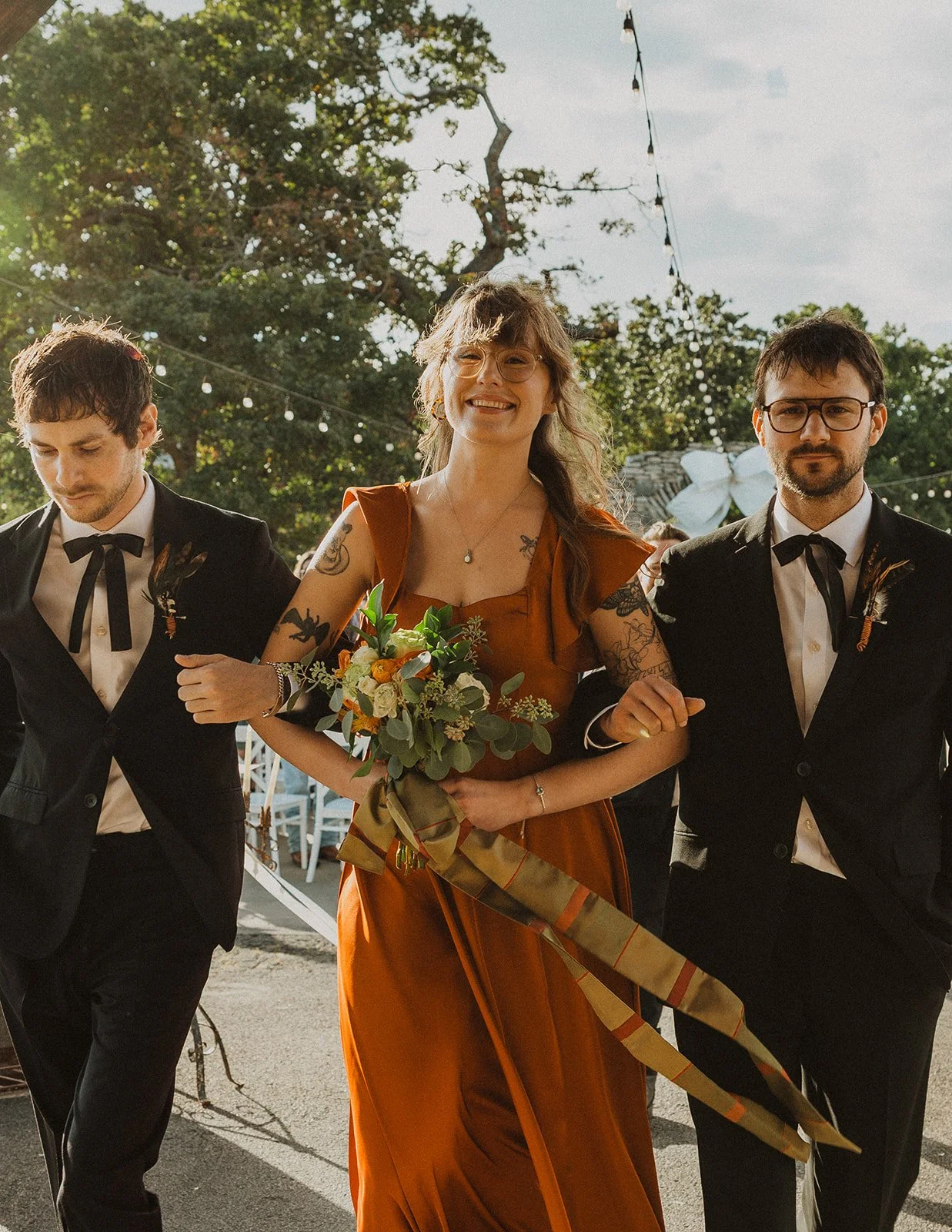 Members of the wedding party walking down the aisle during an outdoor ceremony at Bright Morning Farm with bouquets and black formal attire.