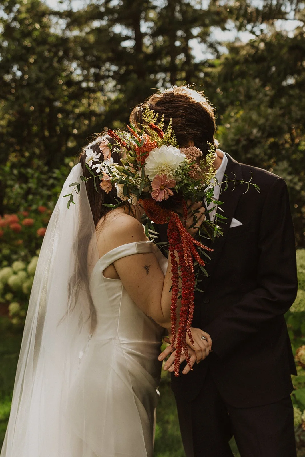 Kissing behind a bouquet of trailing amaranth at a forest wedding