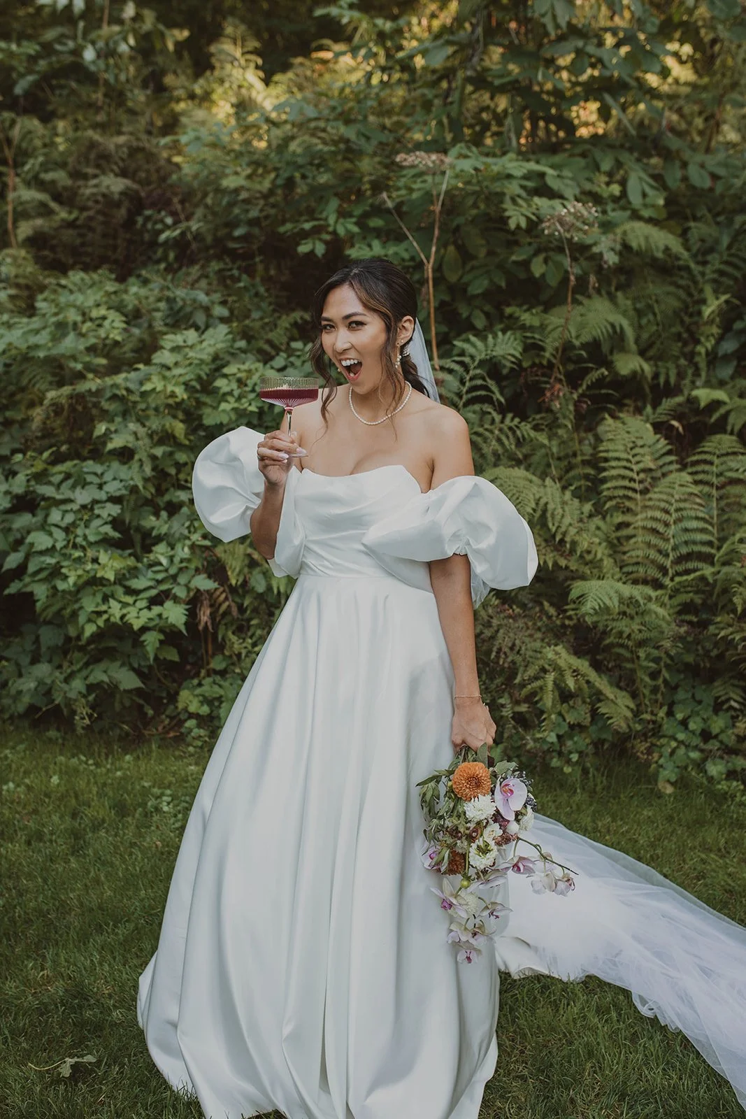 Holding a coupe glass and laughing in front of tall green ferns