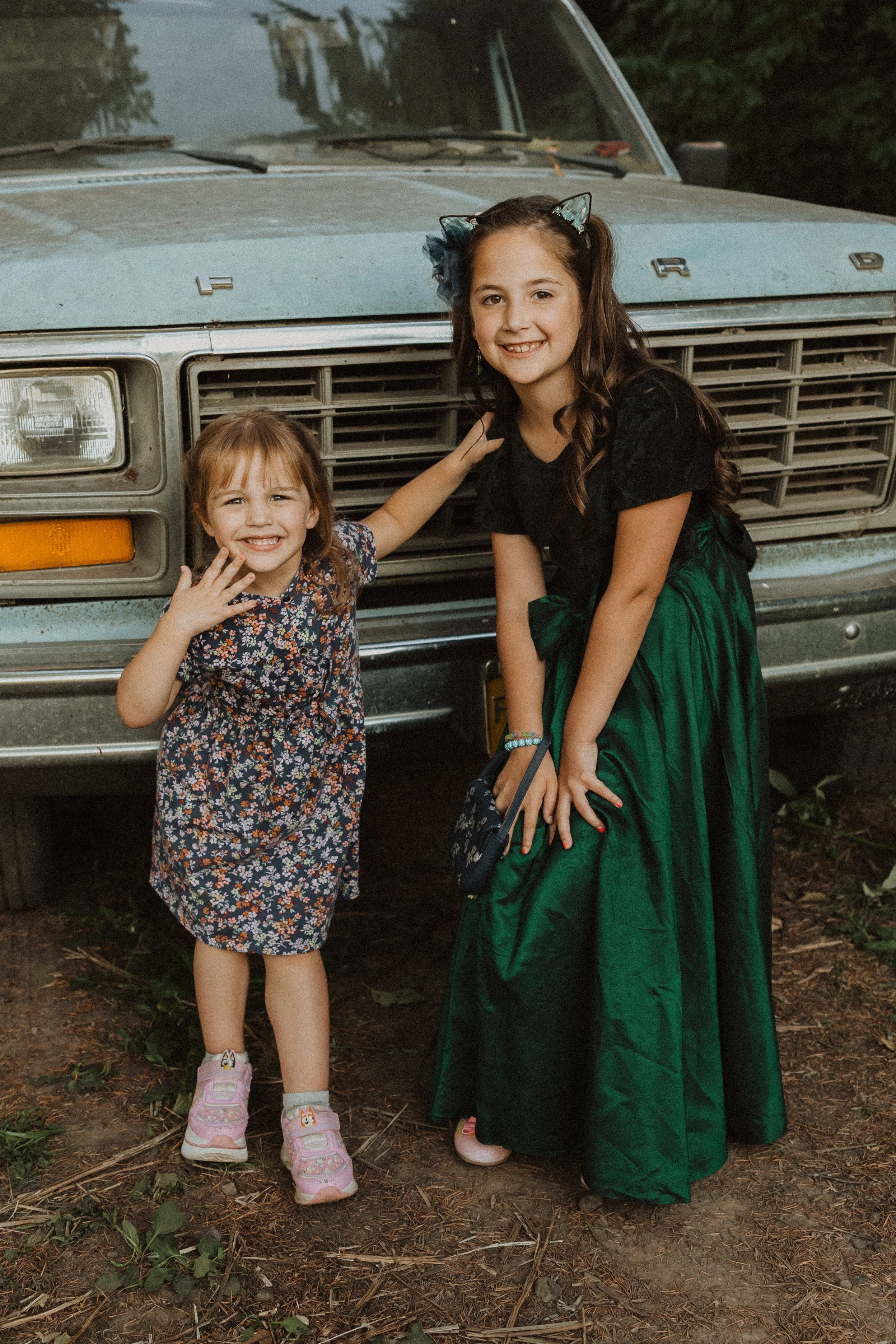 Two smiling girls standing in front of an old, dusty truck, one with her arm around the other. The older girl is wearing a black top and long green skirt, while the younger girl is in a floral dress. They are outdoors on a dirt ground with some green