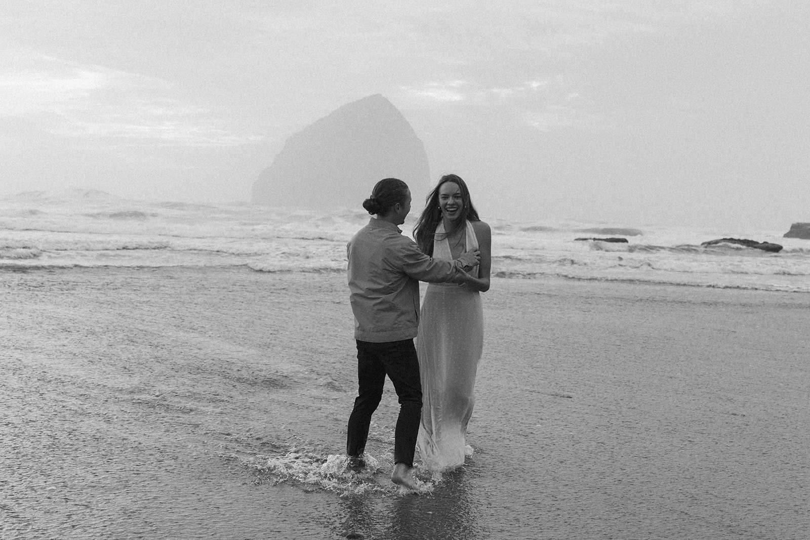 Black-and-white photo of couple laughing together in the shallow water at Cape Kiwanda on the Oregon Coast