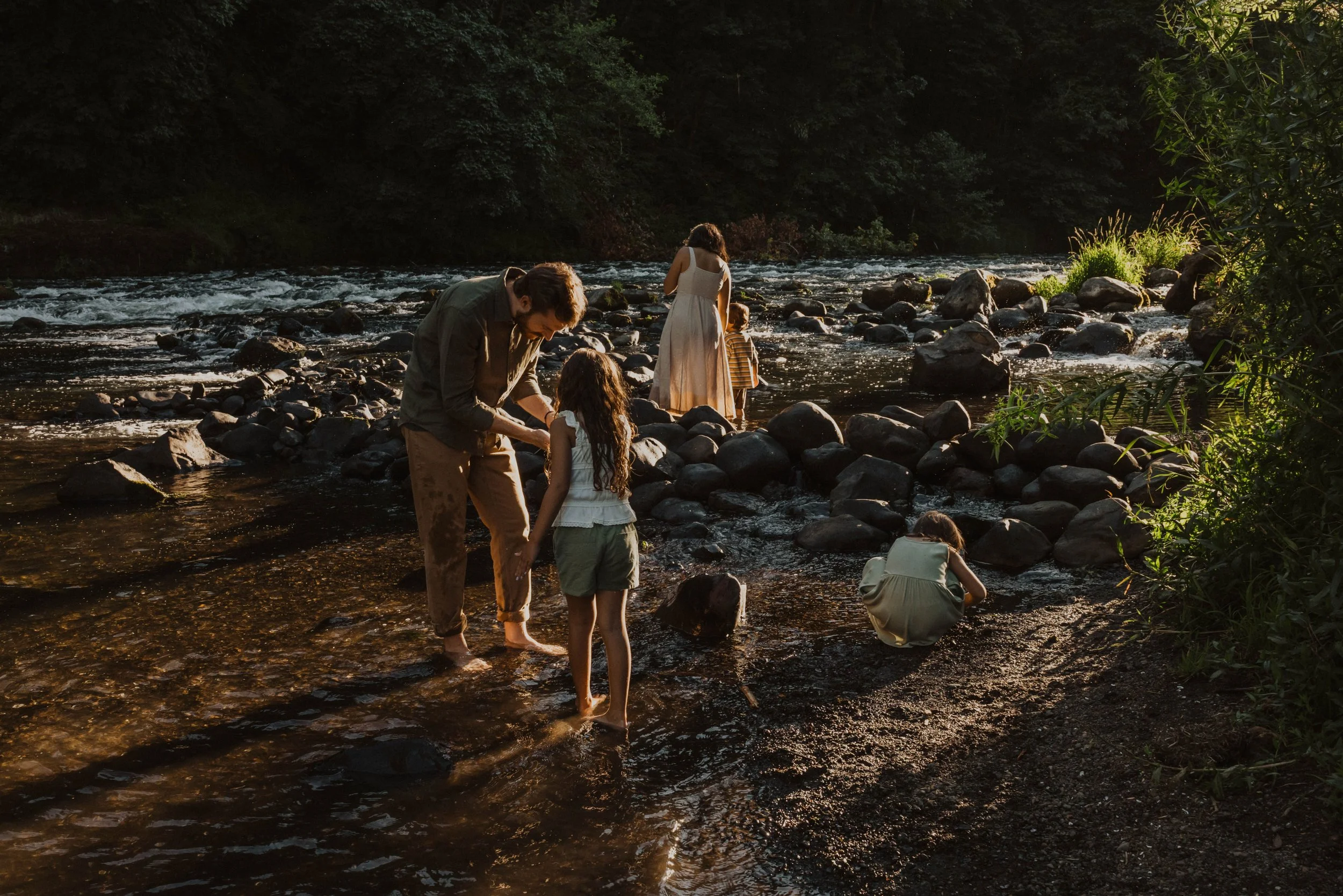 A family of five enjoying a day at a rocky river, with kids playing in the water and adults assisting and watching, surrounded by lush greenery.