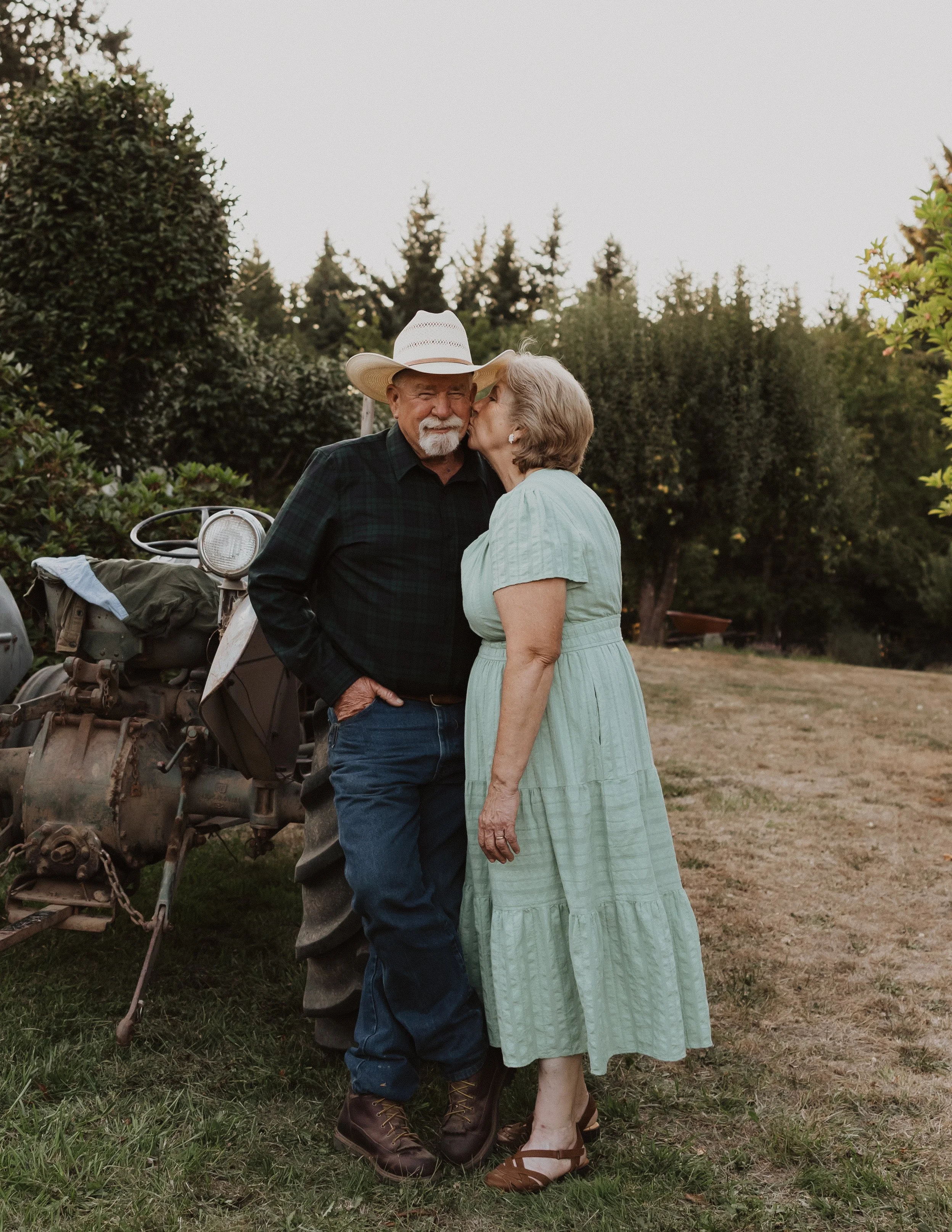 Family Session in Aurora, Oregon. An elderly man in a black shirt, jeans, and a wide-brimmed hat standing close to an elderly woman in a light green dress, sharing a kiss on the cheek outdoors at sunset, with trees and a tractor in the background.
