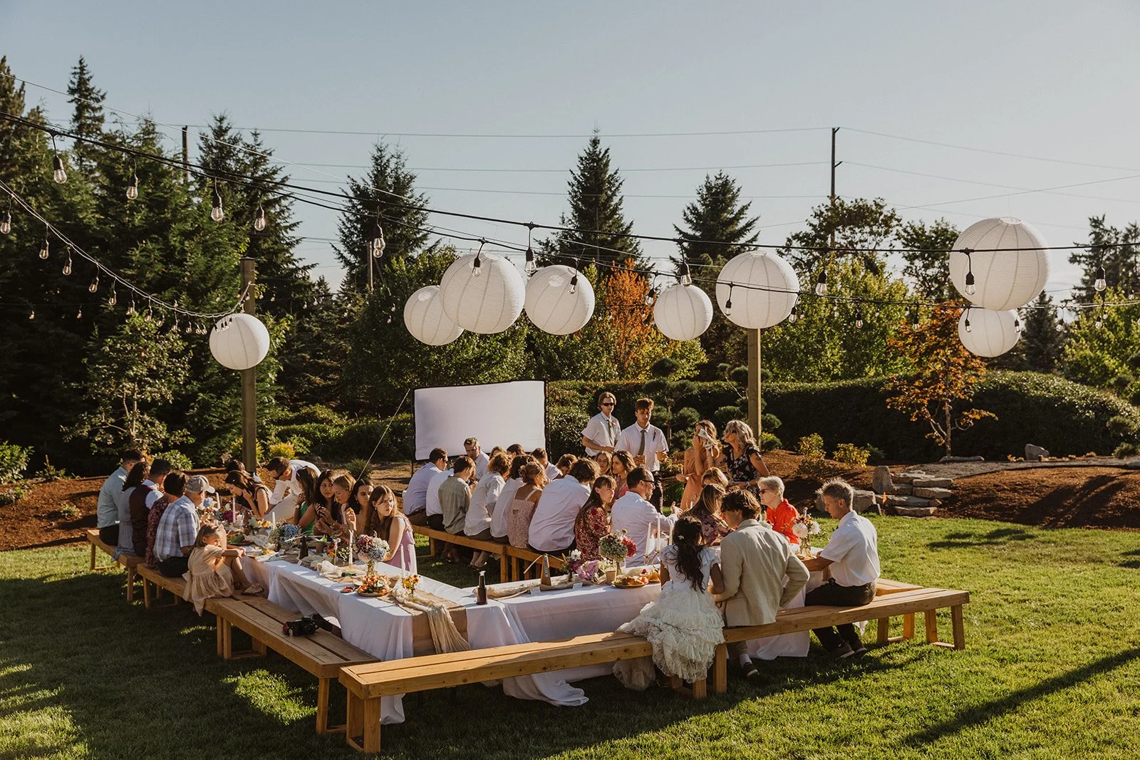 Guests seated at long outdoor reception tables under string lights and white lanterns in a backyard garden setting