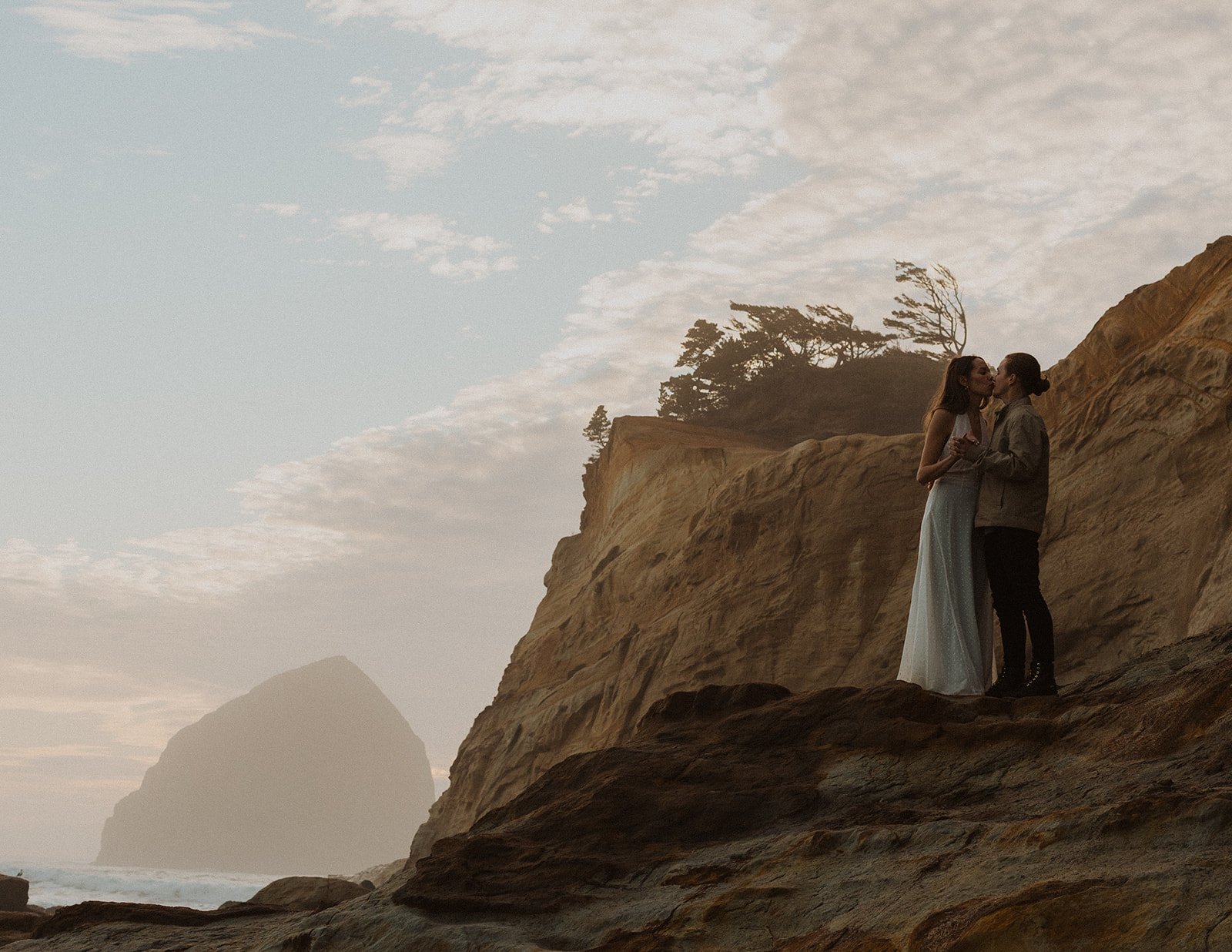Couple standing on coastal cliffs during an Oregon coast engagement session at sunset with ocean and rock formations in the background