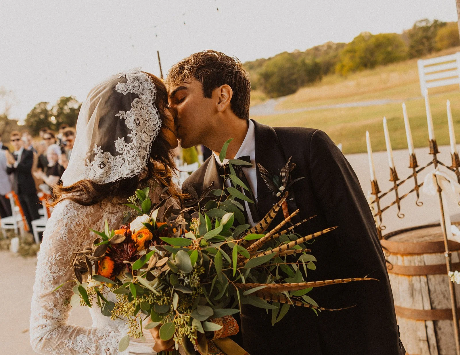 Bride and groom sharing their first kiss during an outdoor fall wedding at Bright Morning Farm near Tulsa, with a large greenery and feather bouquet.