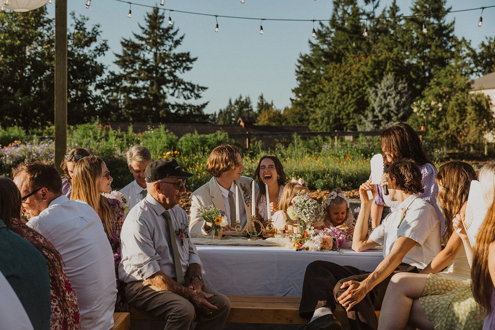 Wedding party and guests seated around a long outdoor reception table, laughing and talking under string lights with flowers and greenery in the background