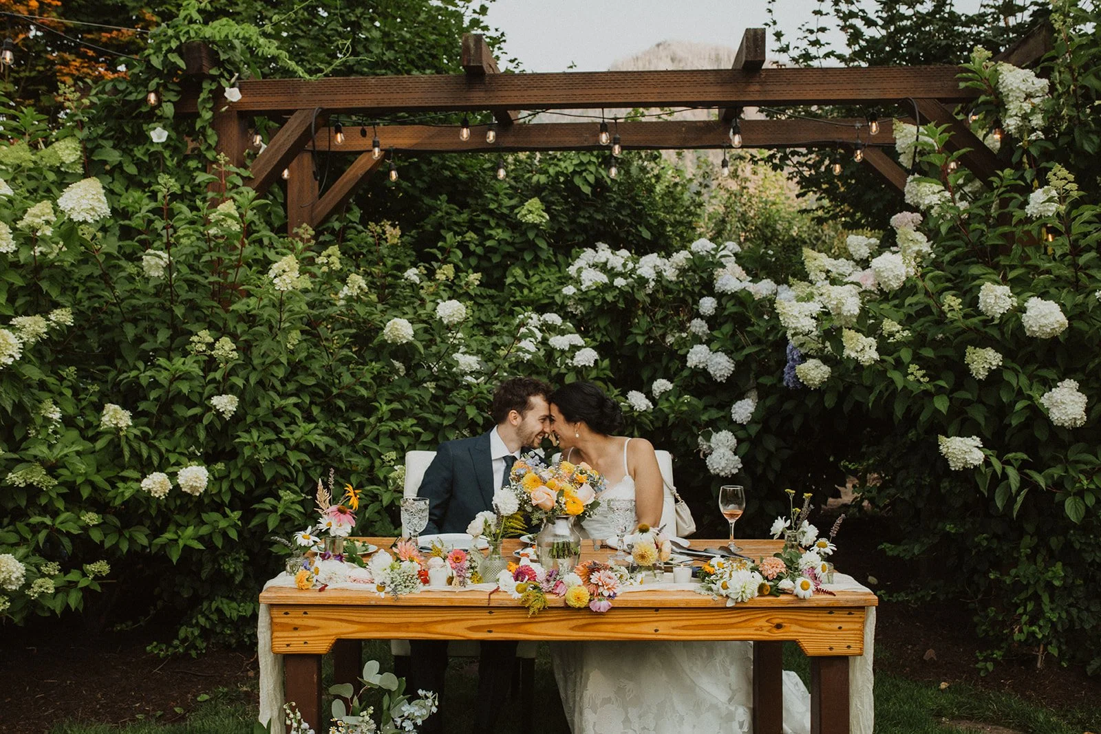 Romantic sweetheart table at an outdoor Cape Horn Estate wedding reception featuring lush hydrangea gardens, string lights, colorful floral arrangements, and the bride and groom sharing an intimate moment together.