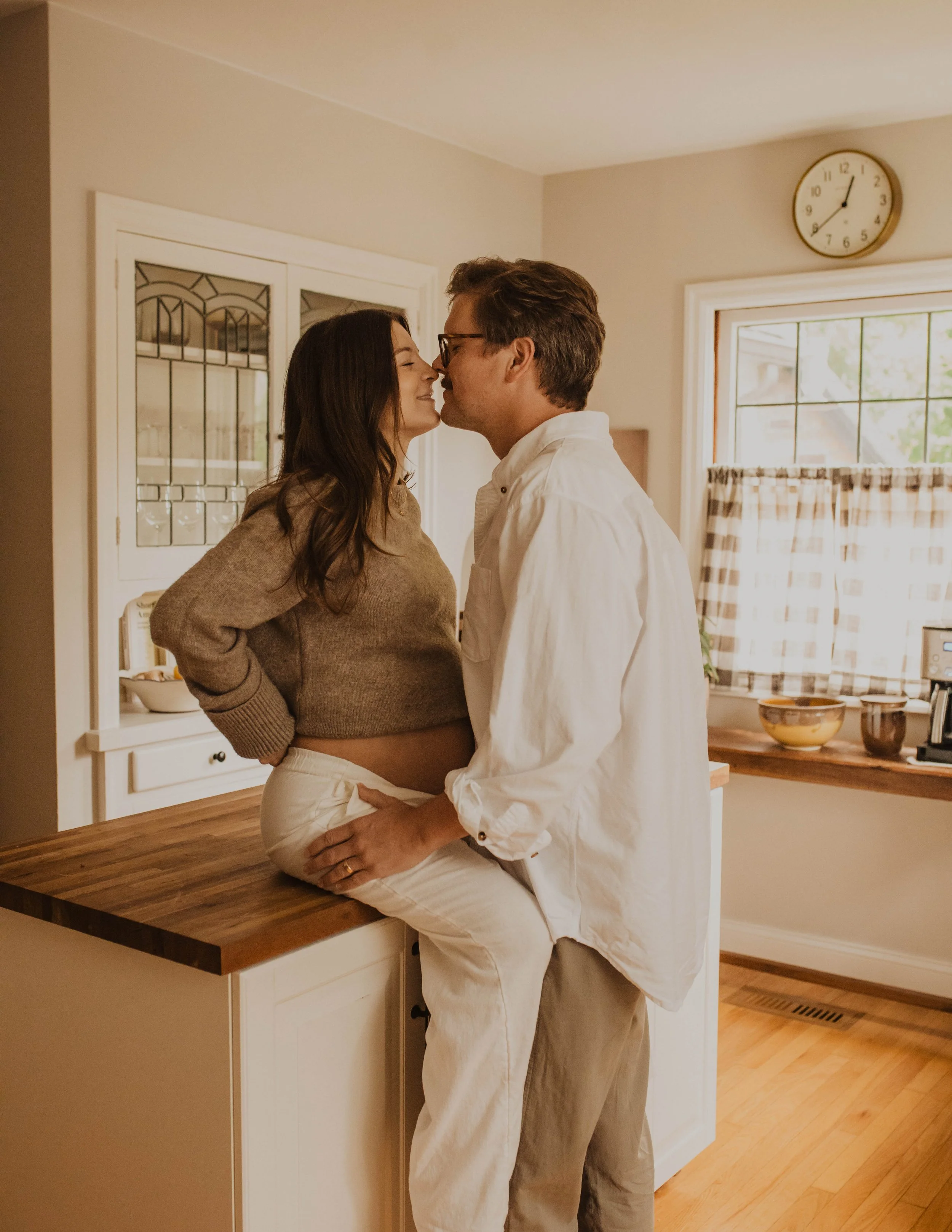 A couple sharing an intimate moment in their kitchen, with the woman sitting on a kitchen counter and the man standing close, touching her thigh as they lean in for a kiss.