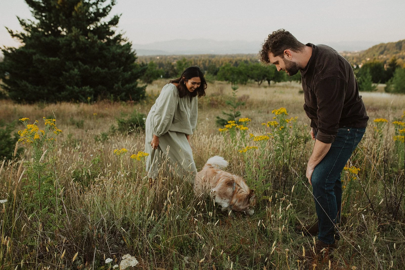 Couple laughing and interacting with their dog in a grassy meadow during a candid Oregon engagement session