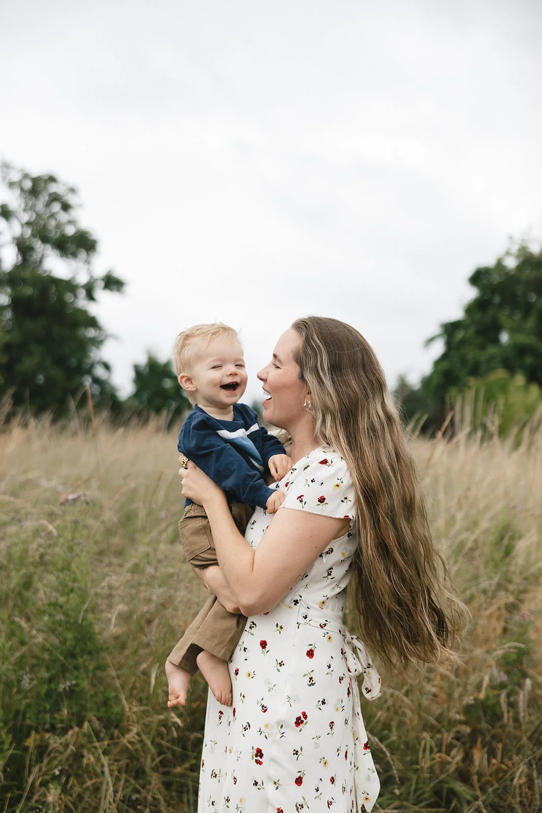 A mom and her two year old son laughing with each other in field of grass on a warm summer morning.