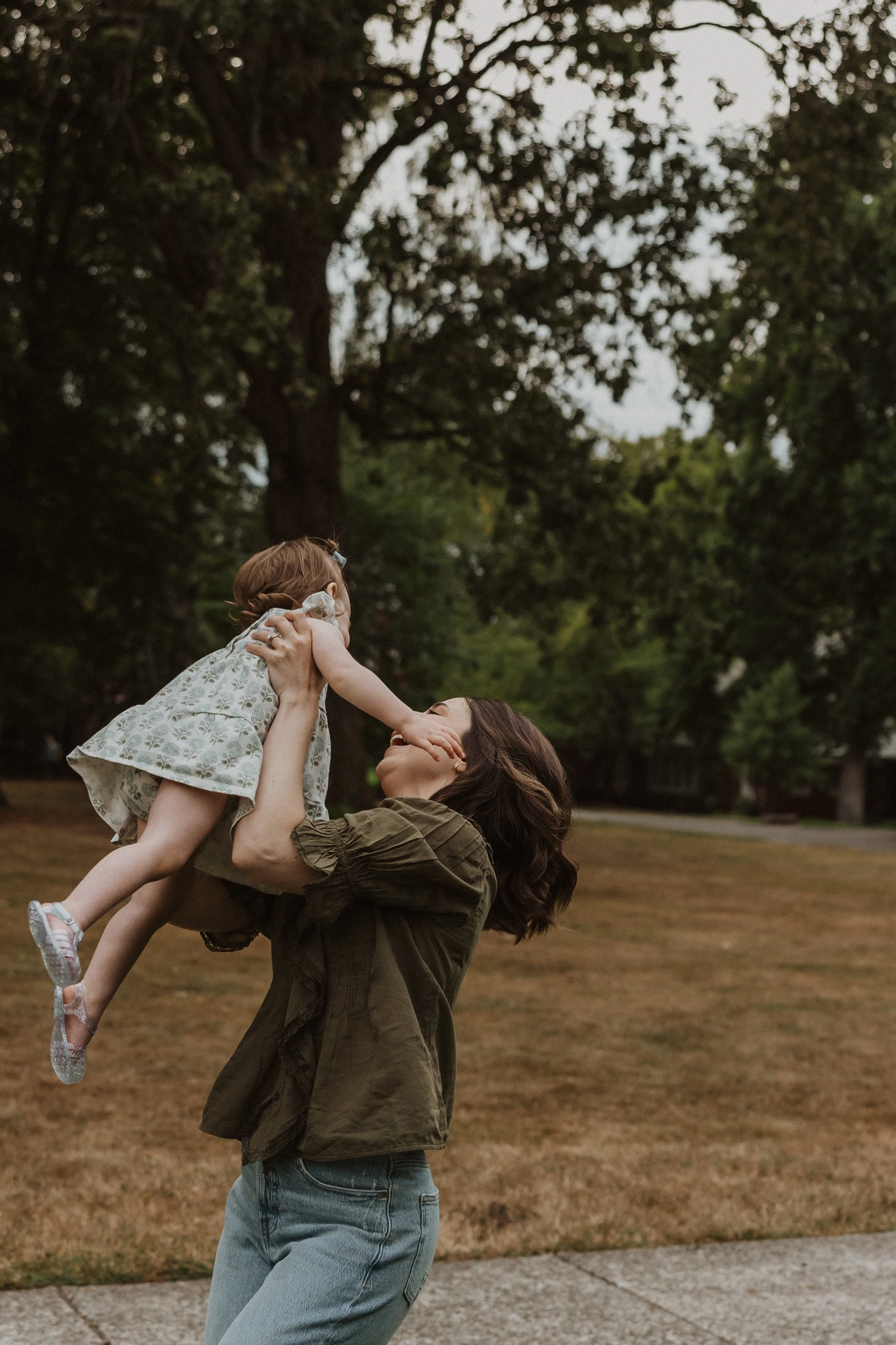 A woman lifting a young girl into the air outdoors in a park with grass and trees.