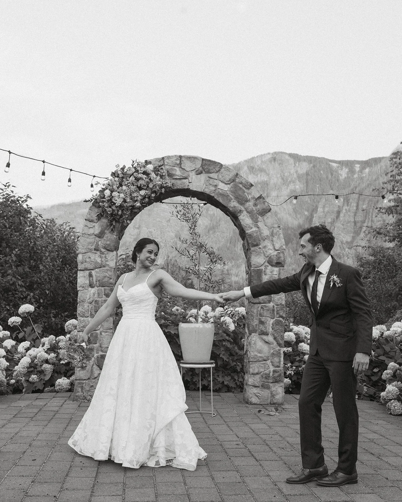 Bride and groom holding hands and playfully twirling beneath a stone ceremony arch with floral arrangements and mountain views in the background during an outdoor wedding.