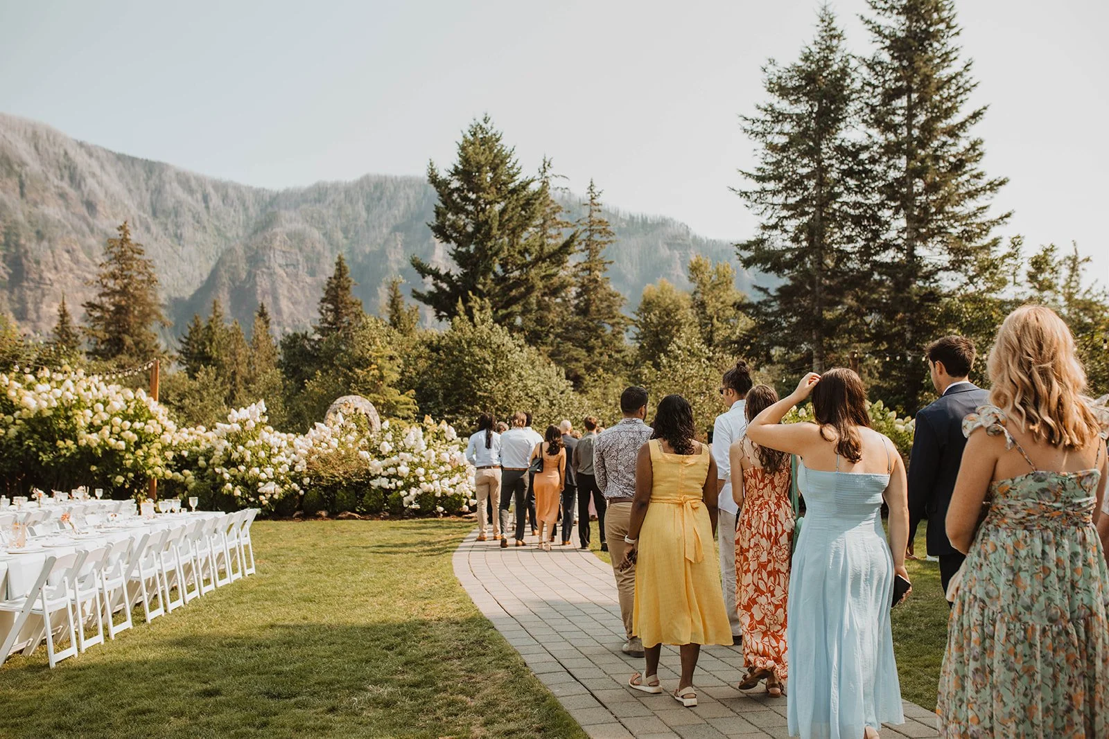 Wedding guests walk along a garden pathway at an outdoor mountain wedding venue surrounded by hydrangeas, long reception tables, and scenic forest views.
