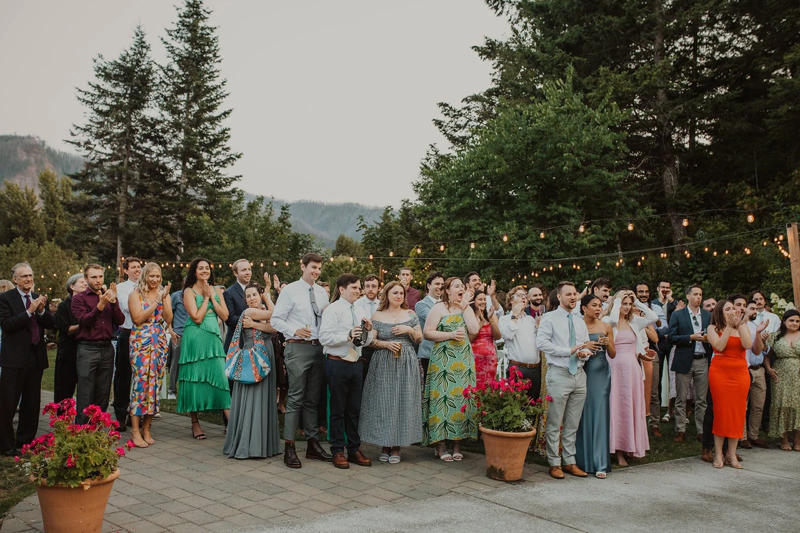 Wedding guests gathered outdoors under string lights at a garden reception venue, smiling and reacting during a speech with mountain views in the background.