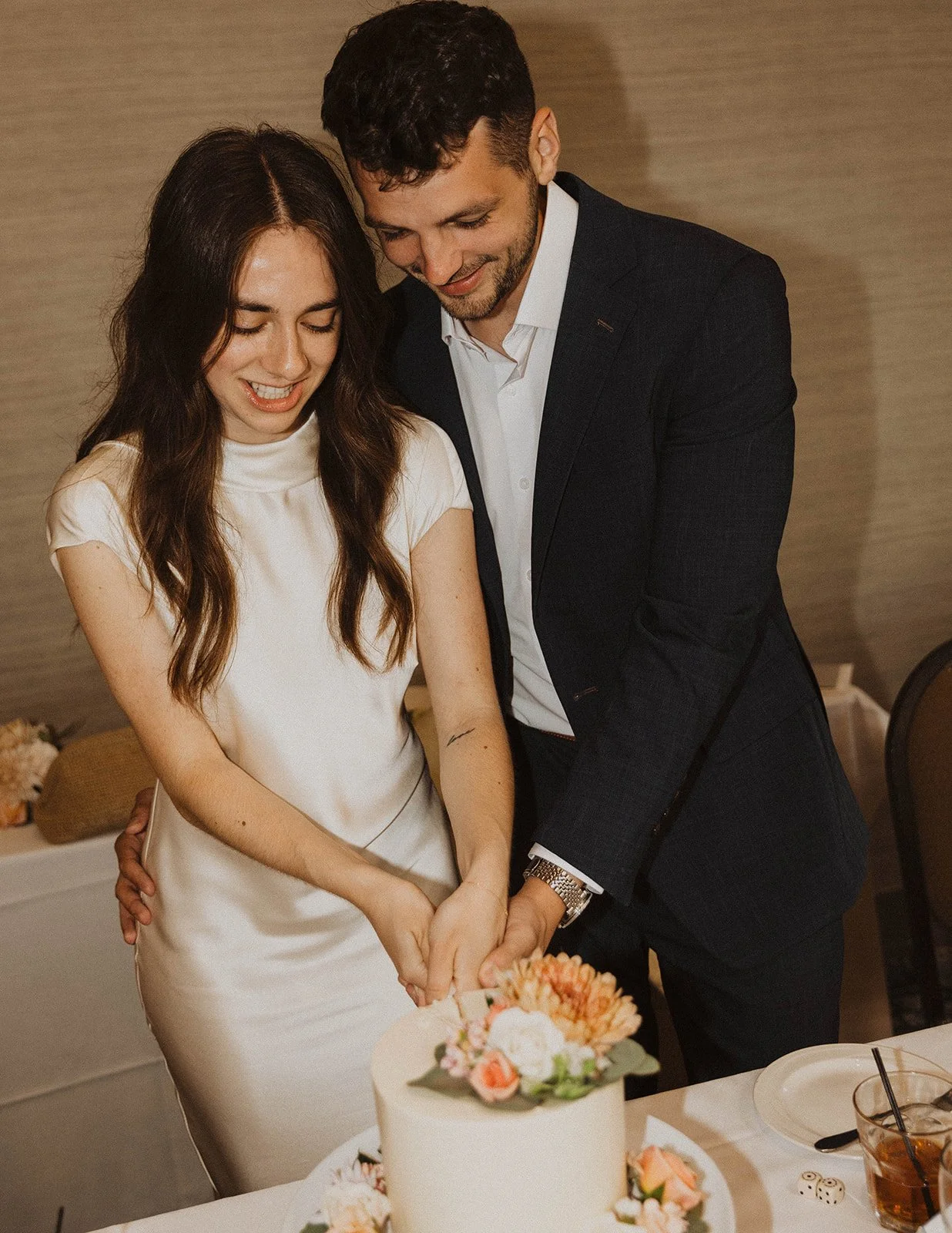 Newlywed couple smiling together while cutting a minimalist floral wedding cake during their intimate reception