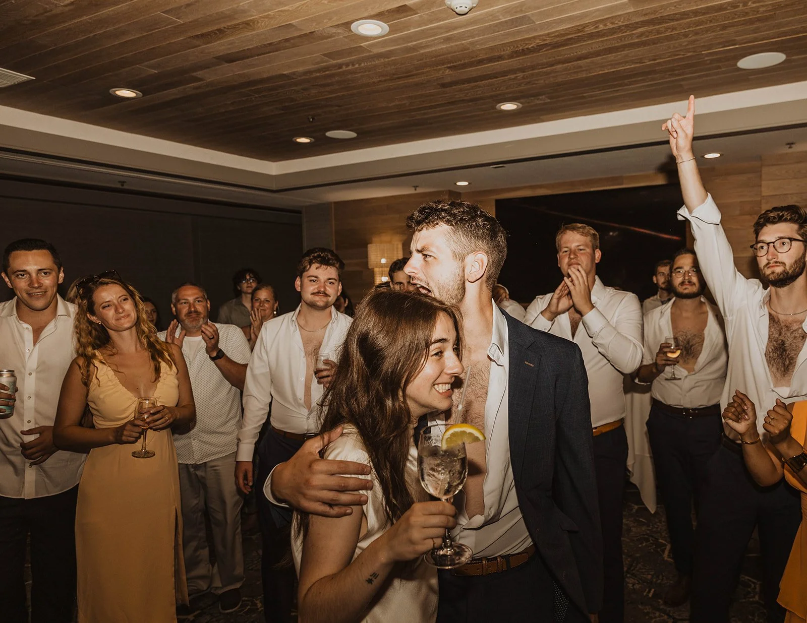 A group of people at a party or celebration, with a focus on a couple dancing in the foreground. The woman is smiling and holding a cocktail, while the man has his arm around her. Others in the background are clapping, smiling, and watching the couple.