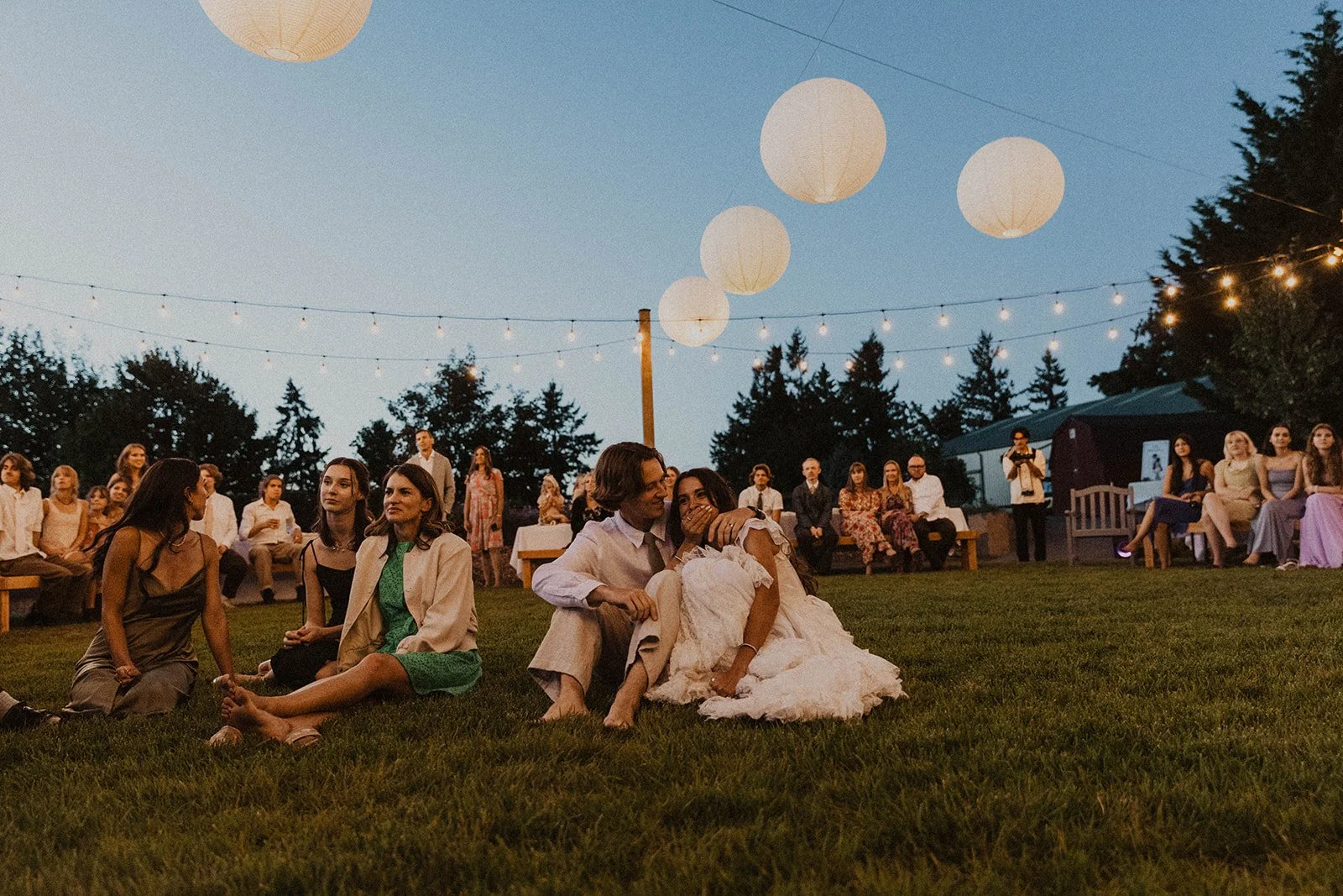 Newlywed couple sitting on the grass embracing while watching a reception moment under string lights with guests in the background