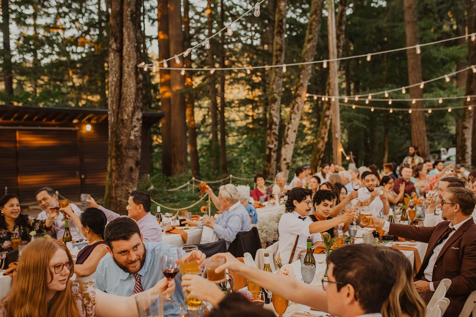 Raising glasses at long tables set among the trees at dusk