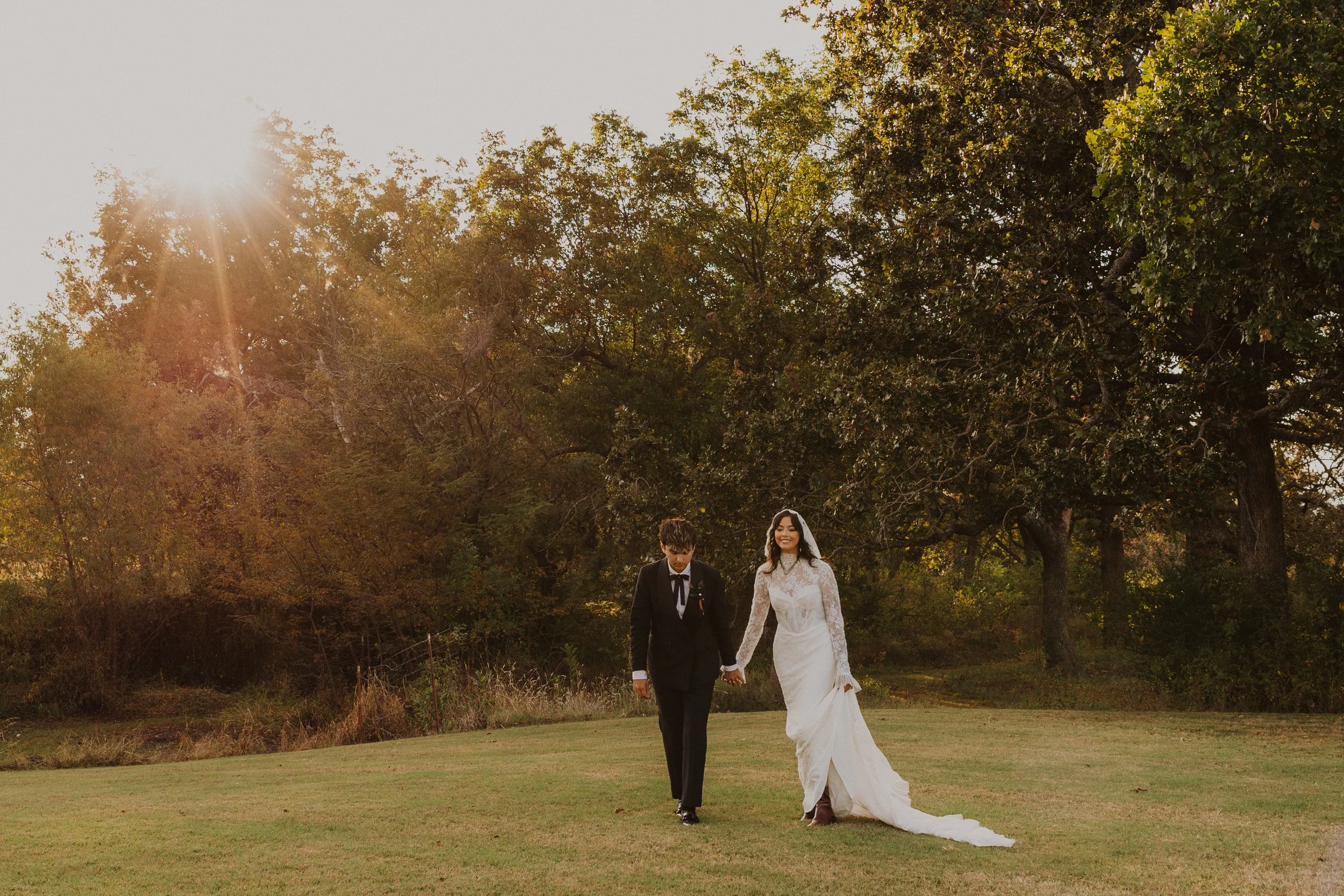 A bride and groom holding hands and walking outdoors on a grassy field with trees in the background during sunset.