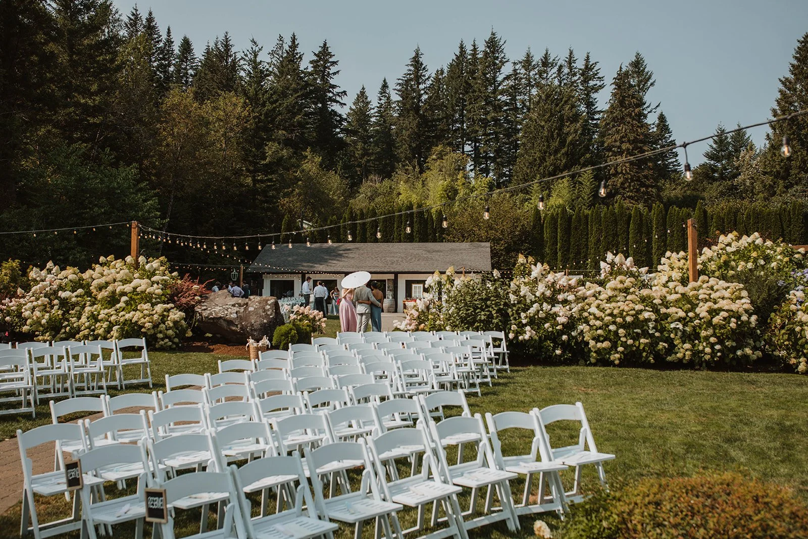 Garden wedding ceremony seating with rows of white chairs, string lights, blooming hydrangeas, and a forested mountain backdrop at an outdoor wedding venue.
