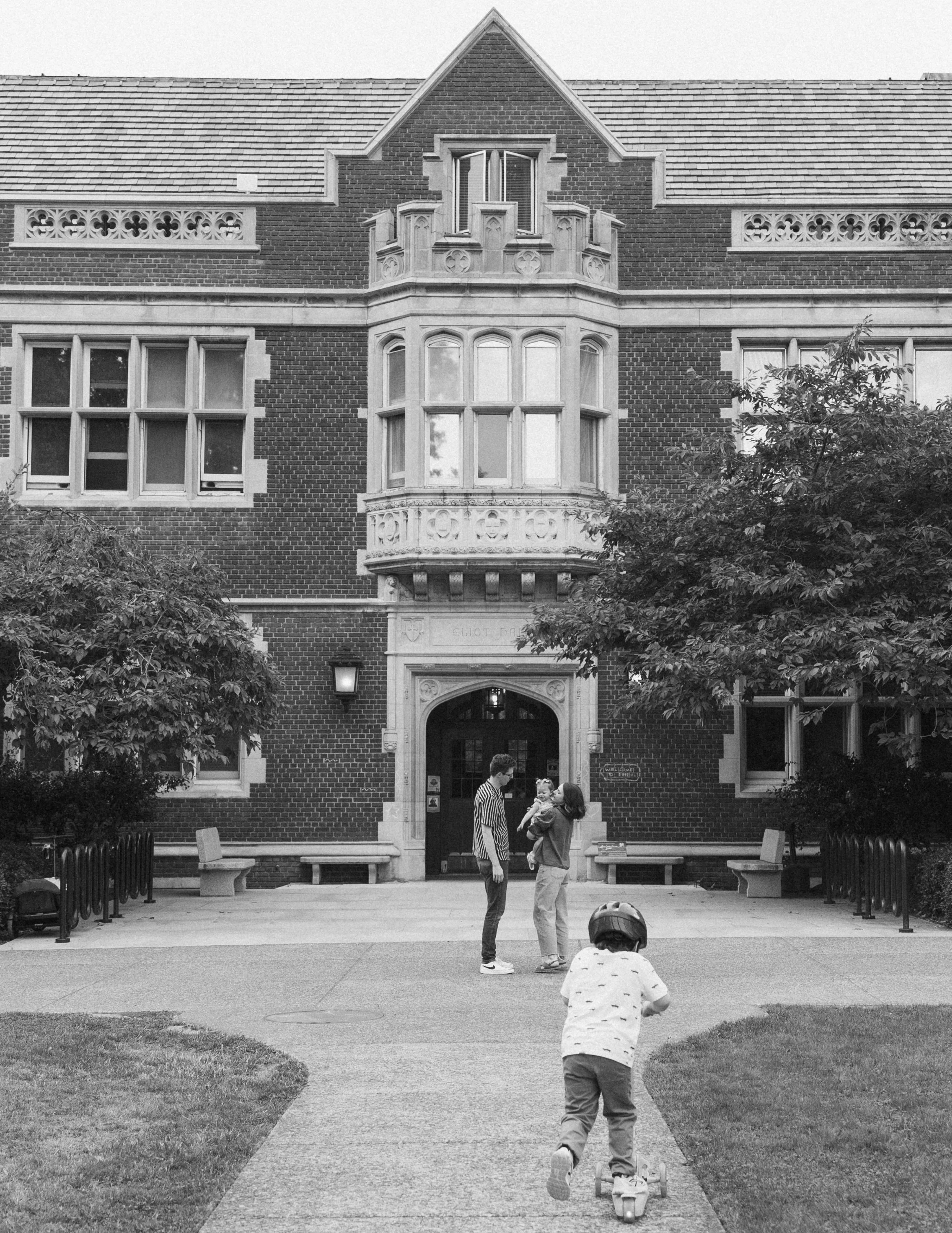 A family in front of a large brick building with a Victorian architectural style, including a bay window and decorative stonework. A person rides a scooter in the foreground, two adults stand talking, and a child is being pulled on a skateboard.