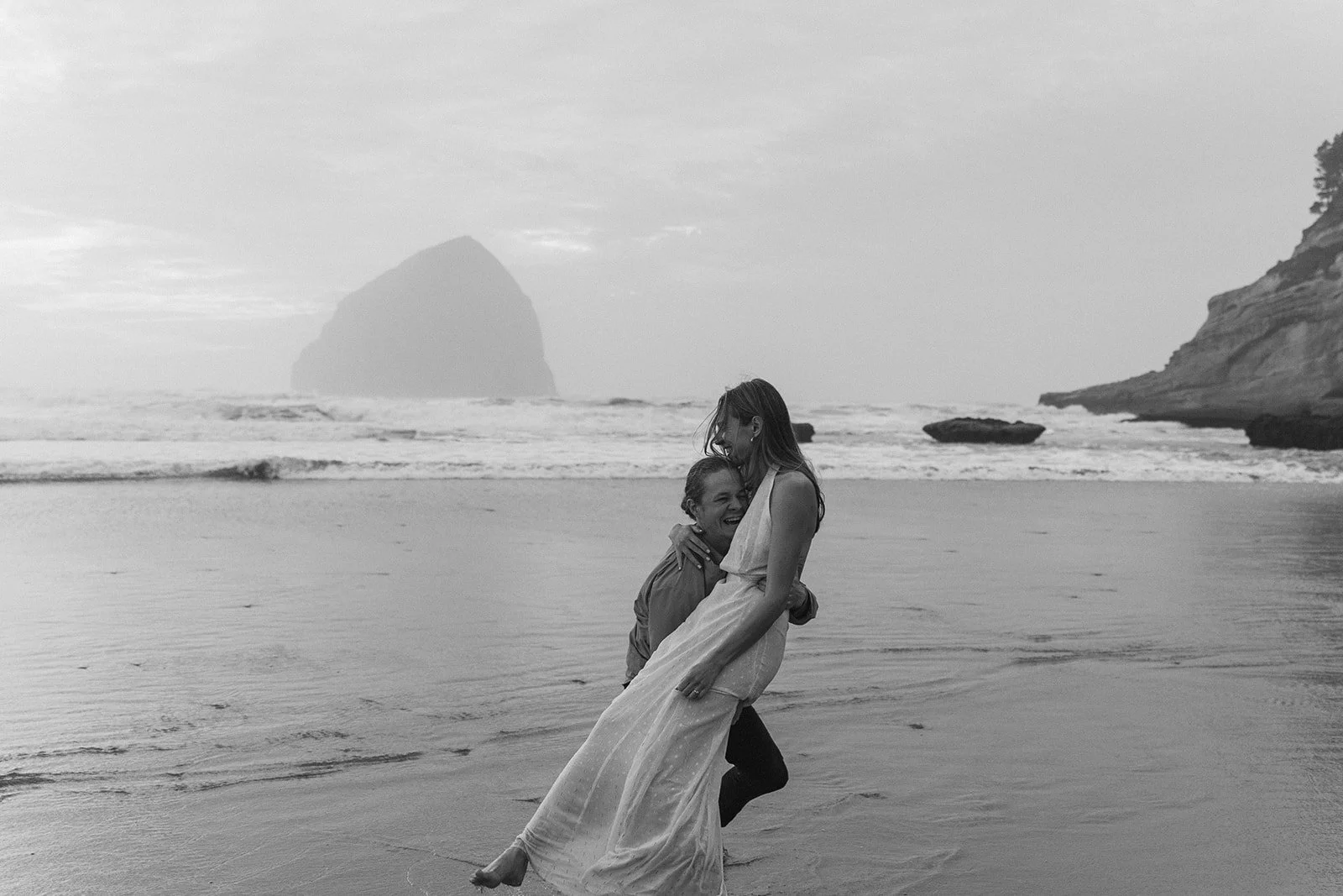 Black and white engagement photo of a couple laughing as he lifts her on the Oregon coast beach with ocean waves behind them