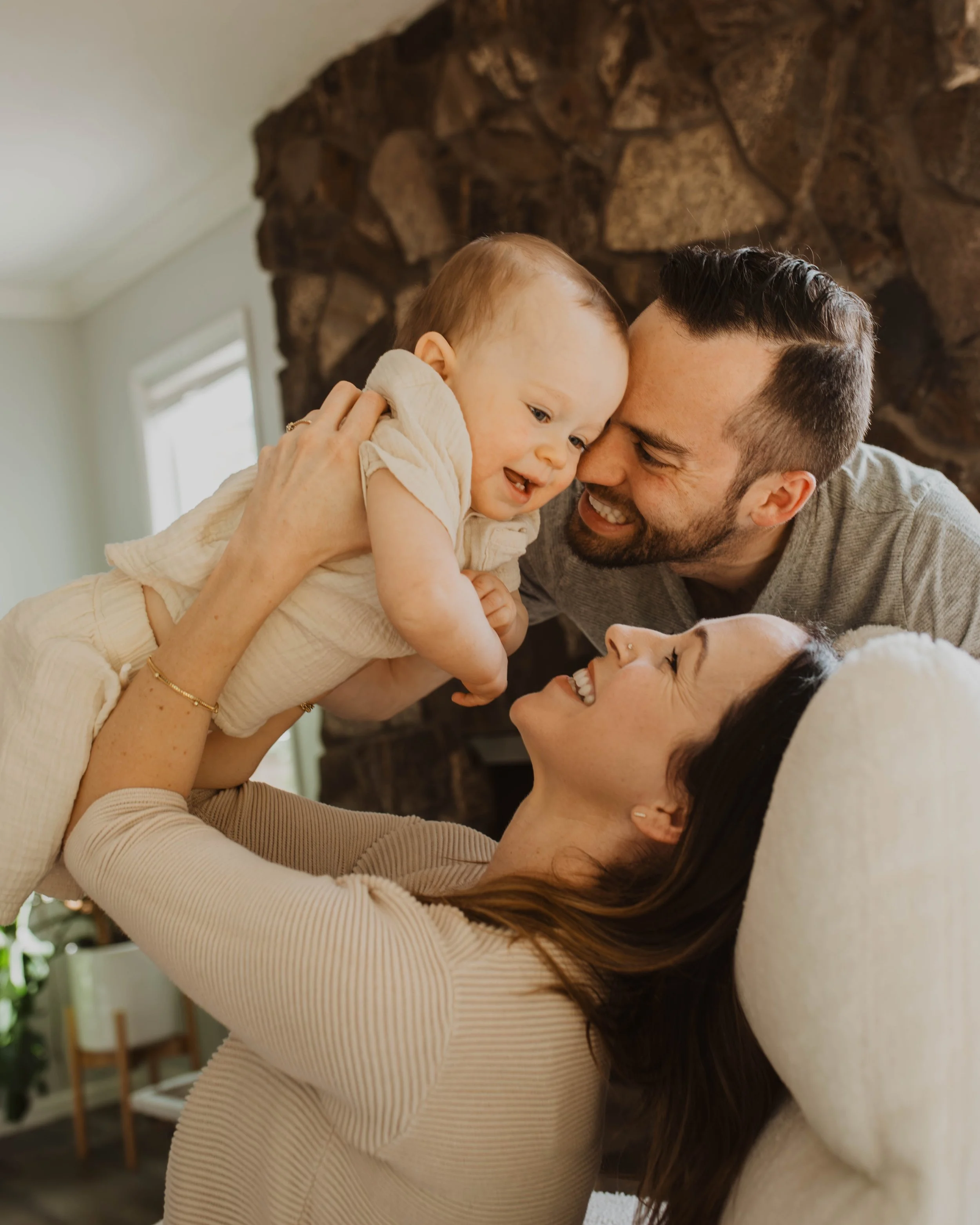 Happy family playing together in a cozy living room: a woman lying on a couch, holding a young child, and a man leaning in to join, all smiling and enjoying a joyful moment.