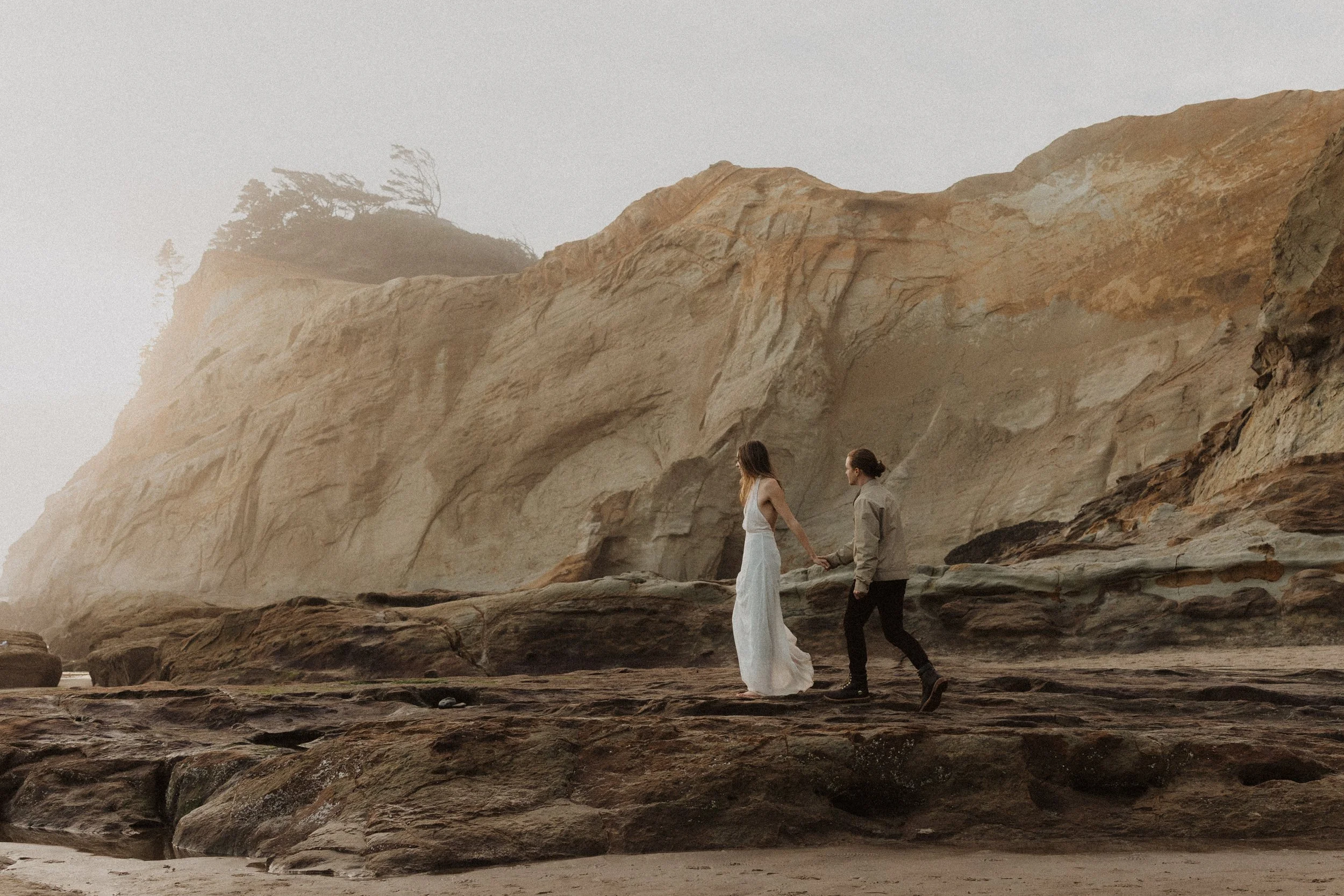 A couple holding hands and walking on a rocky beach with large cliffs and a tree-topped hill in the background, atmospheric and foggy.