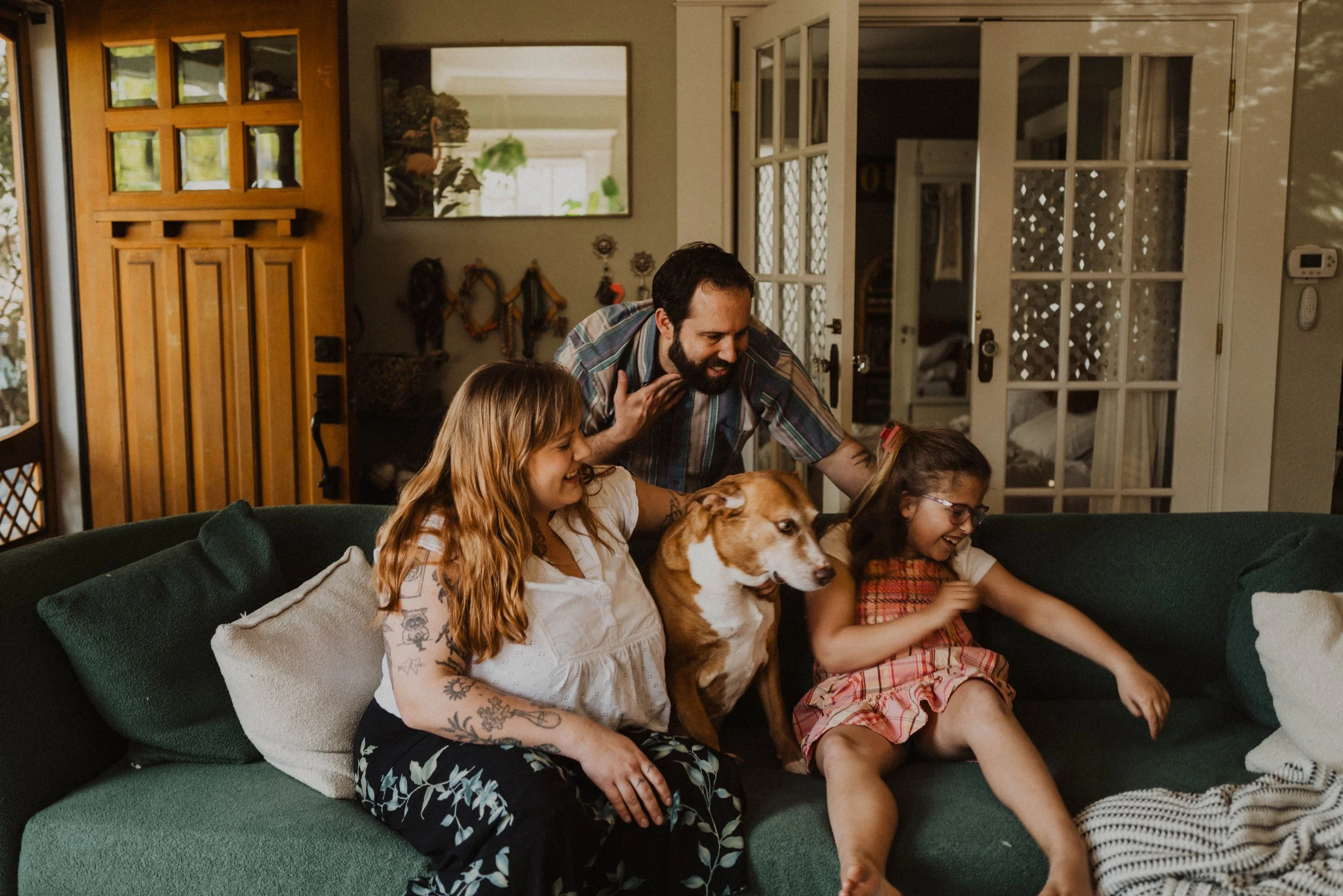 A family of four, including two young girls and a man, sitting on a green couch in a living room, with a brown and white dog between the girls. The family is playing and laughing together.