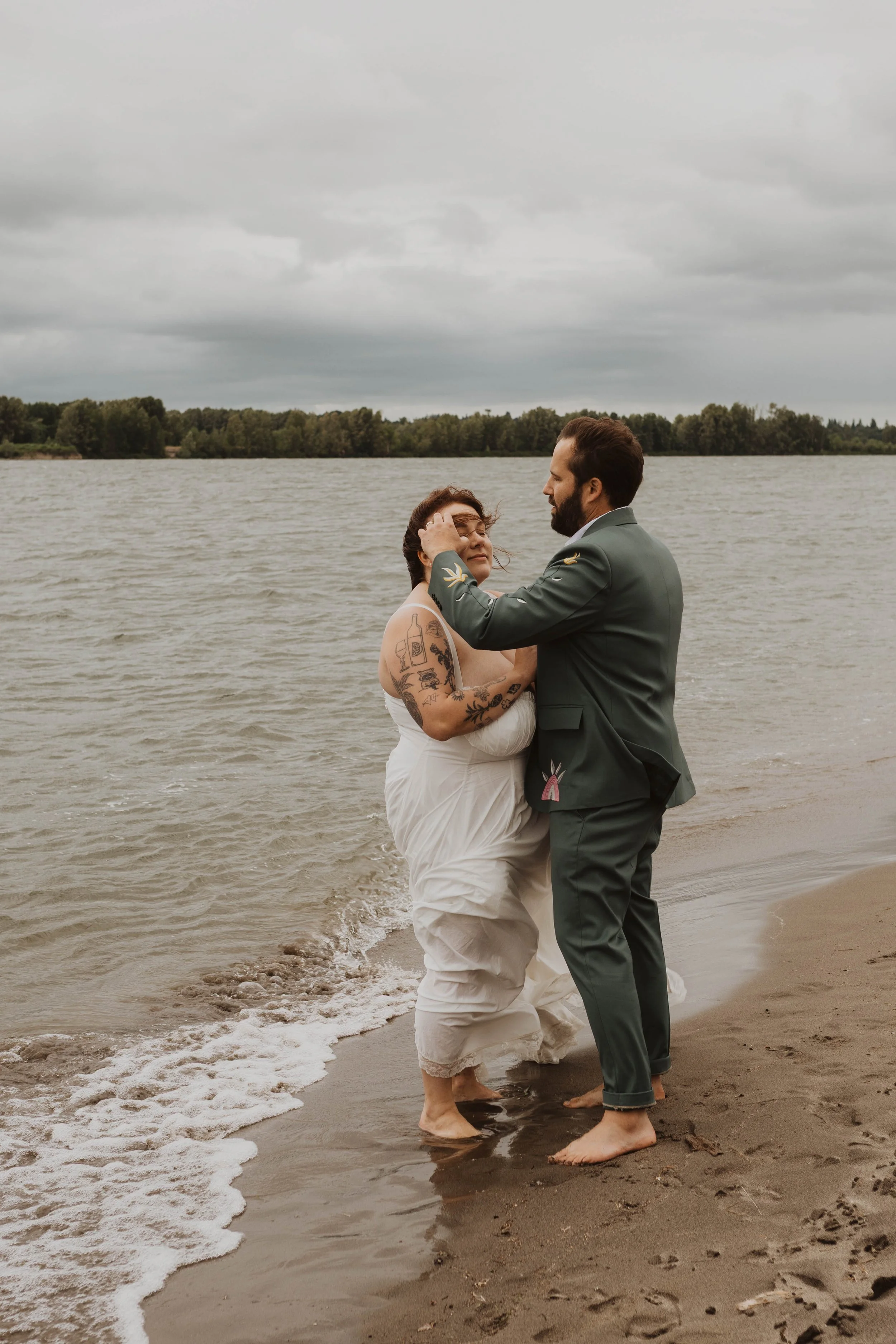A couple in wedding attire standing barefoot on a sandy beach near a lake, embracing each other with overcast skies in the background.