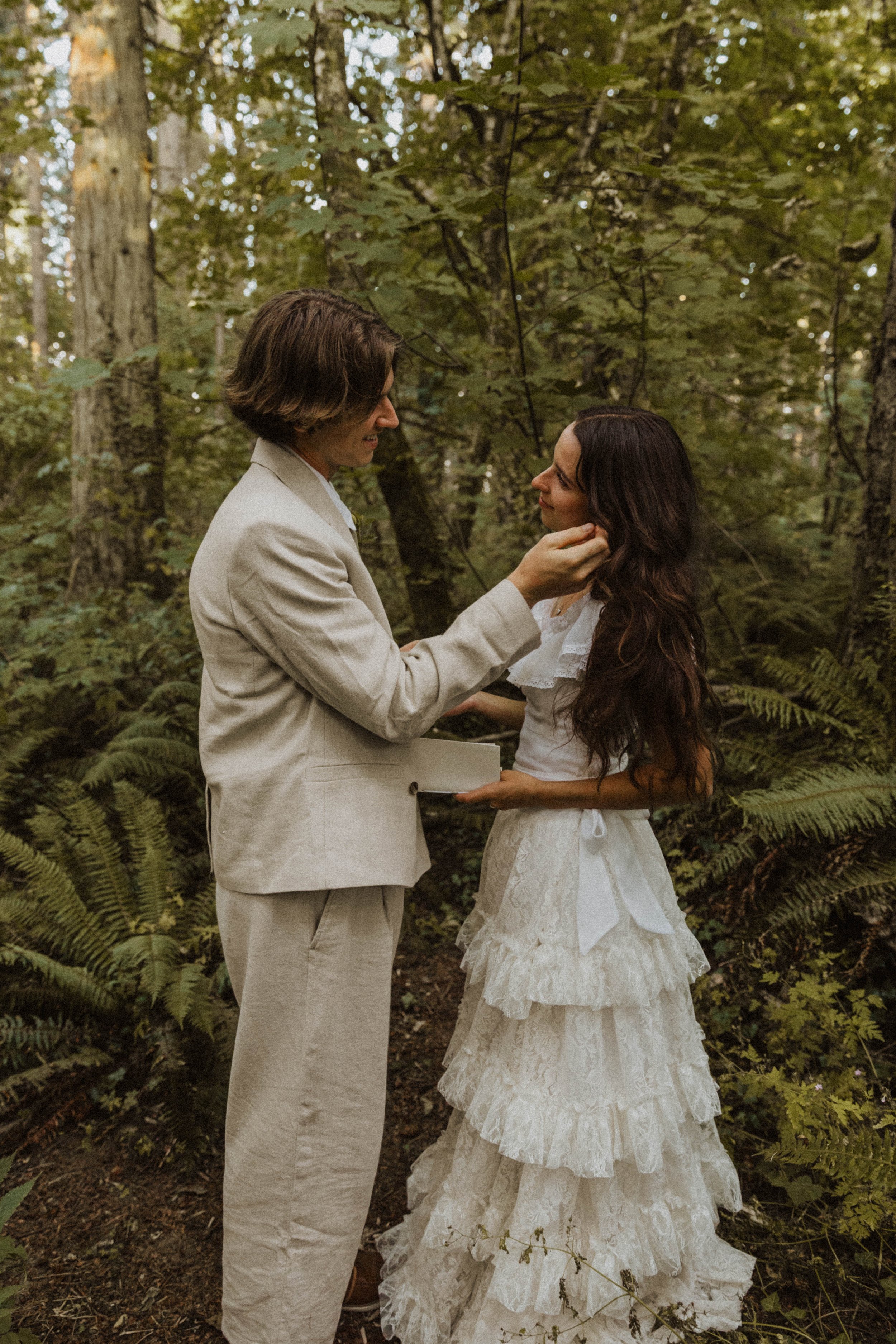 Couple sharing an intimate moment in a lush forest, adjusting hair and smiling softly, captured by an Oregon wedding photographer with a warm, earthy style.